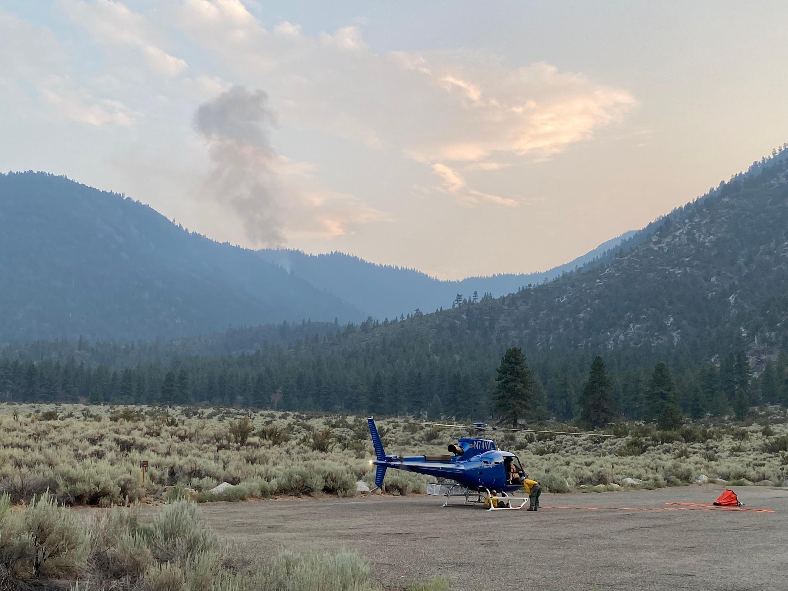 A Bridgeport Helitack crew member loads the A-Star A wildland firefighter ensures the orange water bucket is secure on the helicopter while a smoke column burns in the background