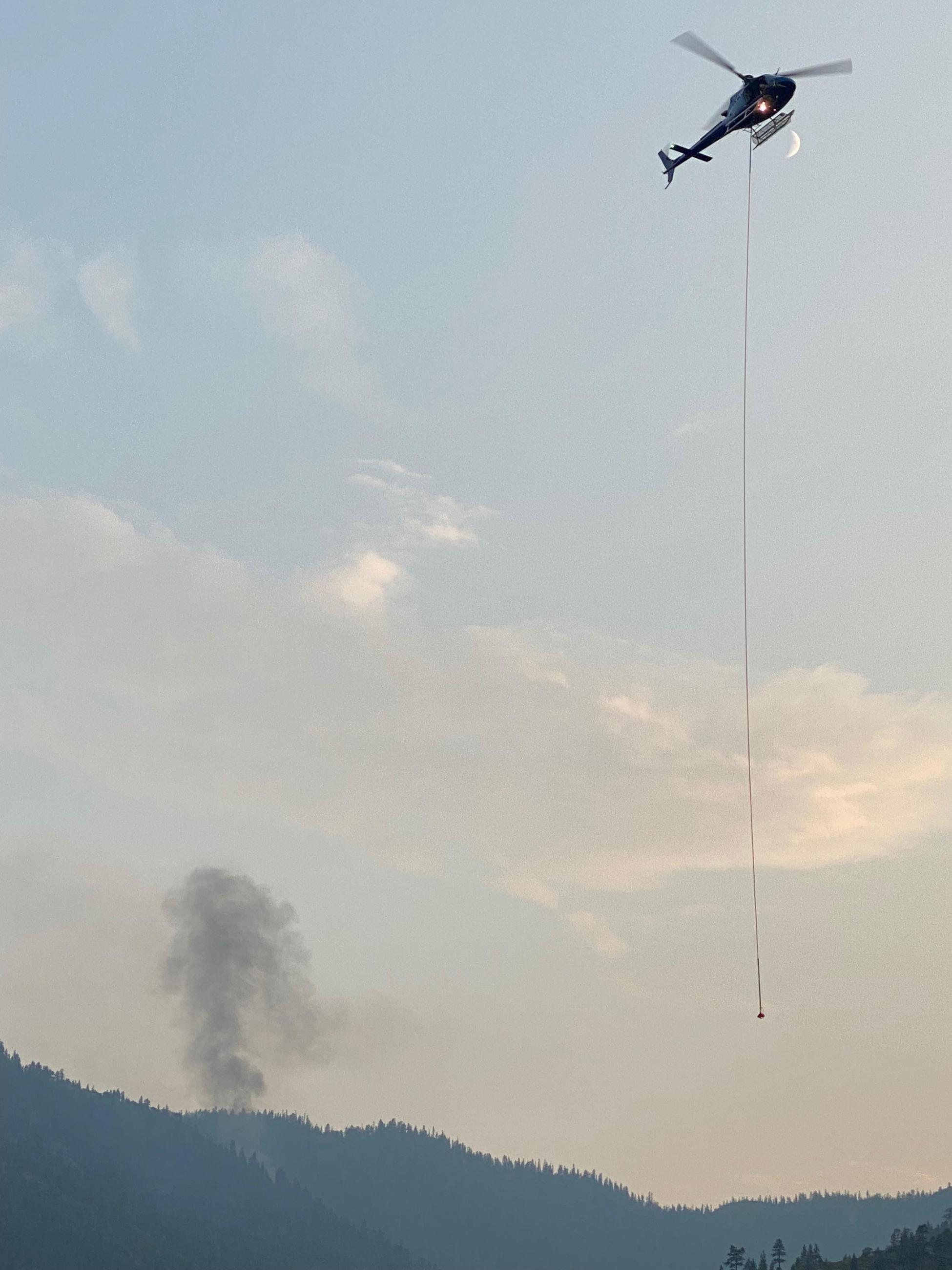 A helicopter with longline flies from the Fay Fire A helicopter with a longline dangling from underneath flies in front of a smoke column emitted from the Fay Fire. A sliver of moon is also visible just below the helicopter.