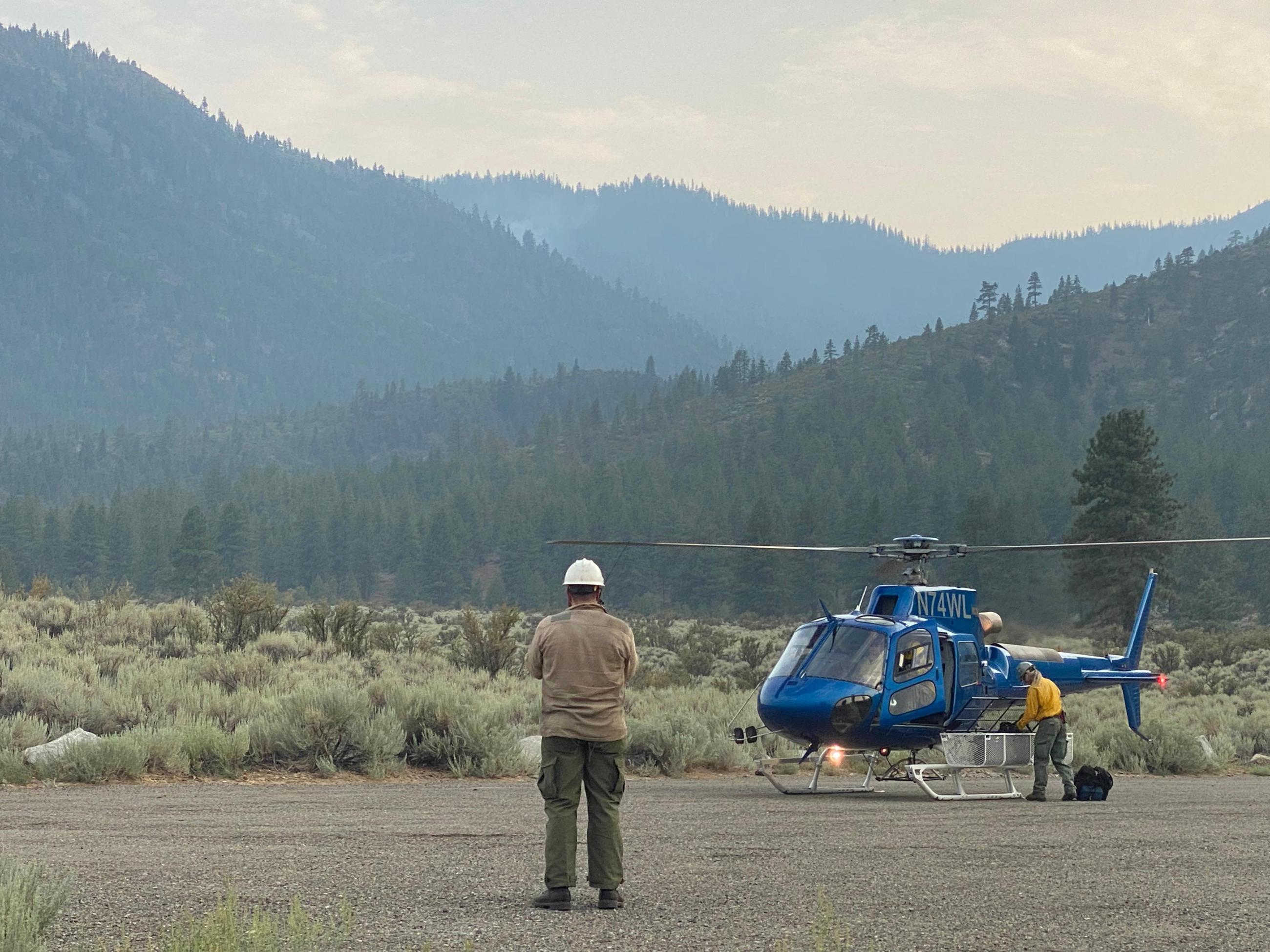 Firefighters carry gear to the helicopter. The mountains in the background are steep and rugged. A firefighter in a white hard hat talks on the radio to the helicopter pilot