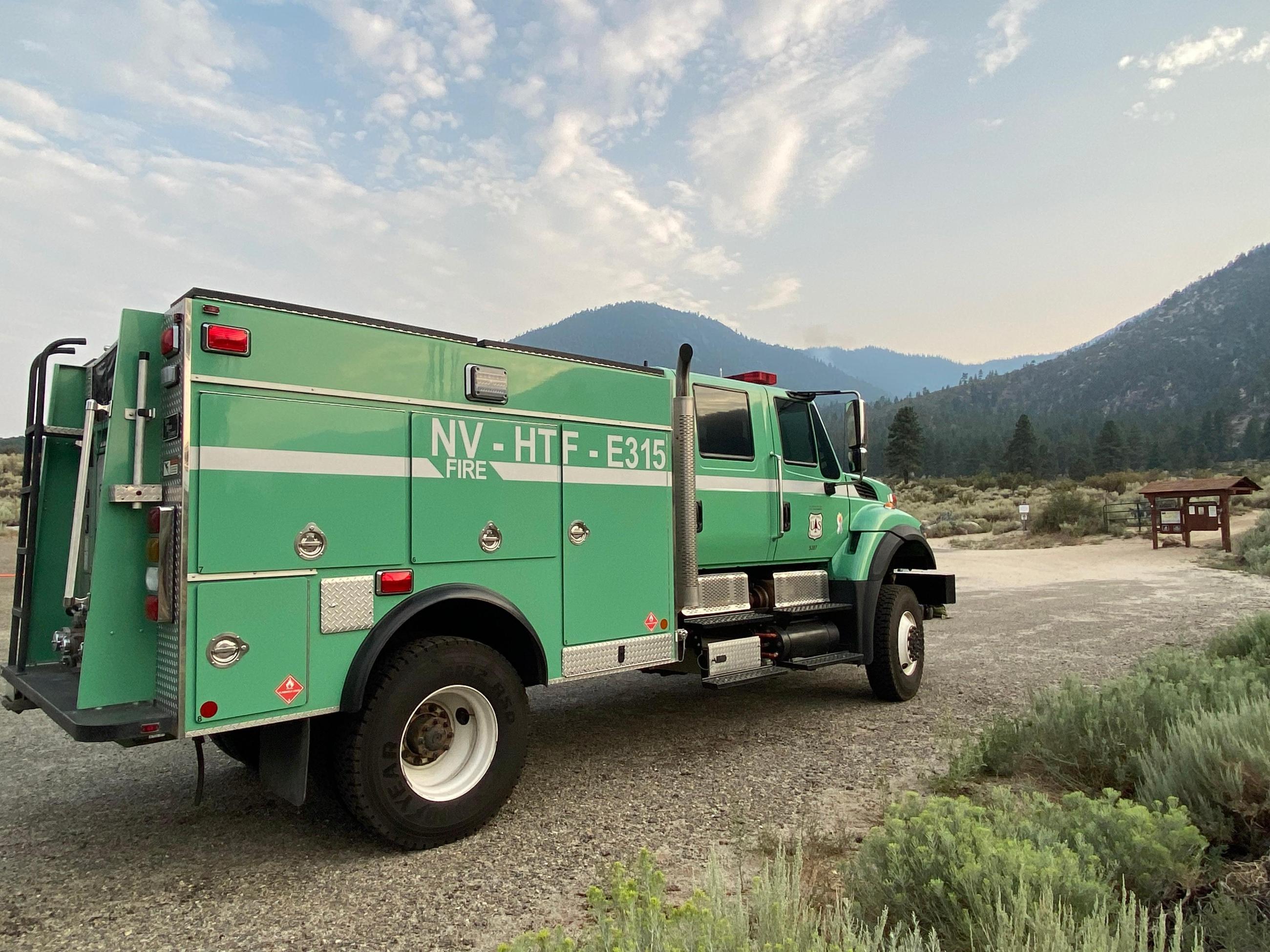 Engine 315 is parked at the trailhead A large, green wildland fire engine with the call sign NV-HTF 315 is parked at a trailhead with mountains and a small, wispy smoke column in the backgournd.
