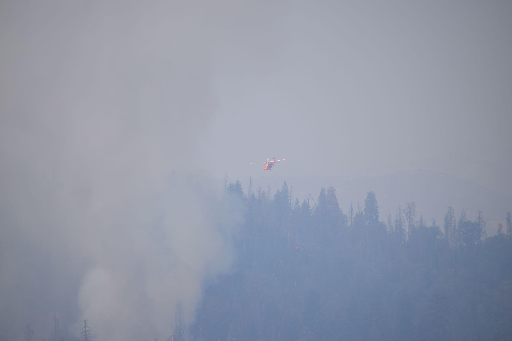 A red helicopter carries a water drop bucket over a very smoky hillside