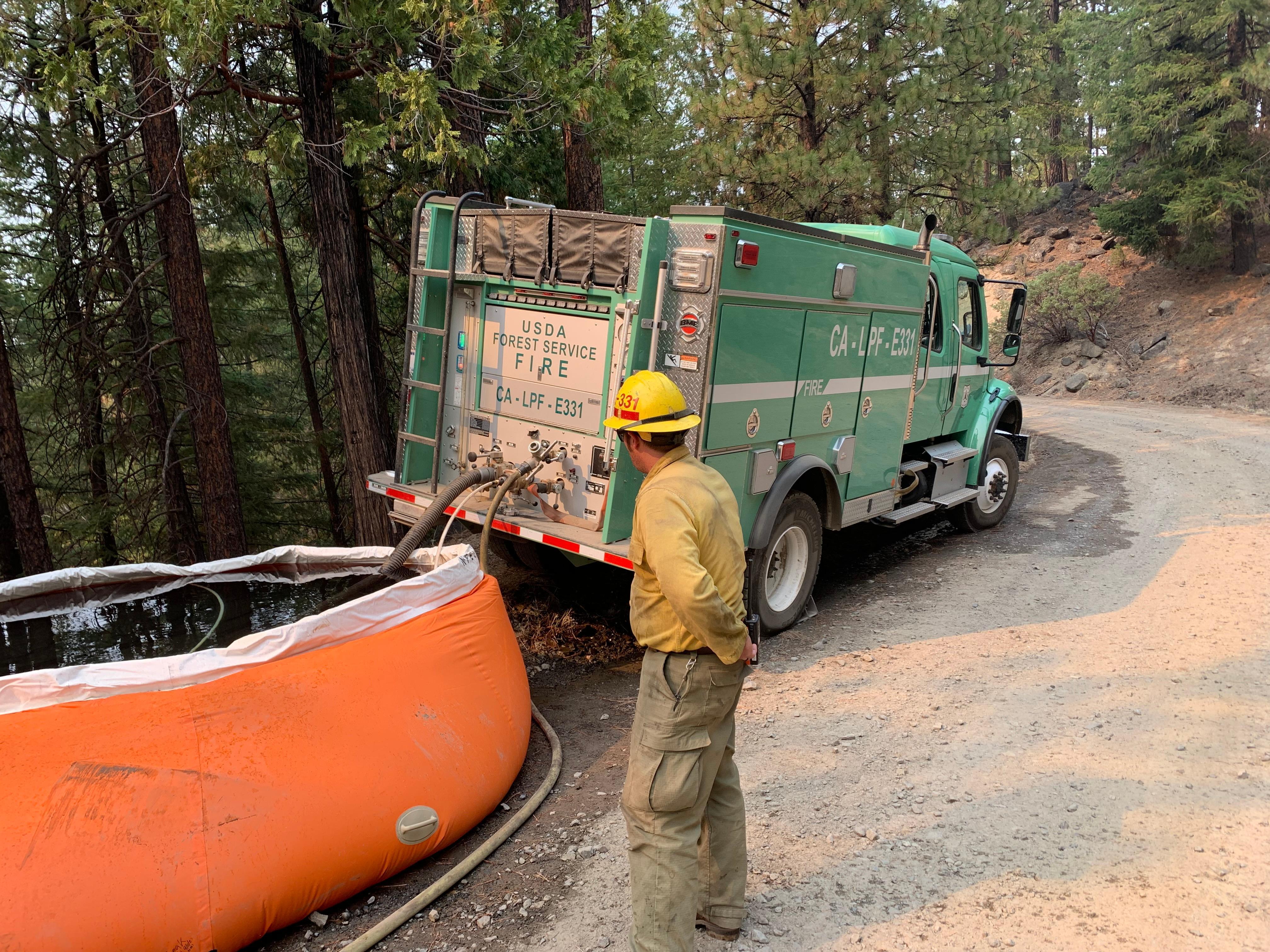 A firefighter looks at a water container and a fire engine on a forested, dirt road. 