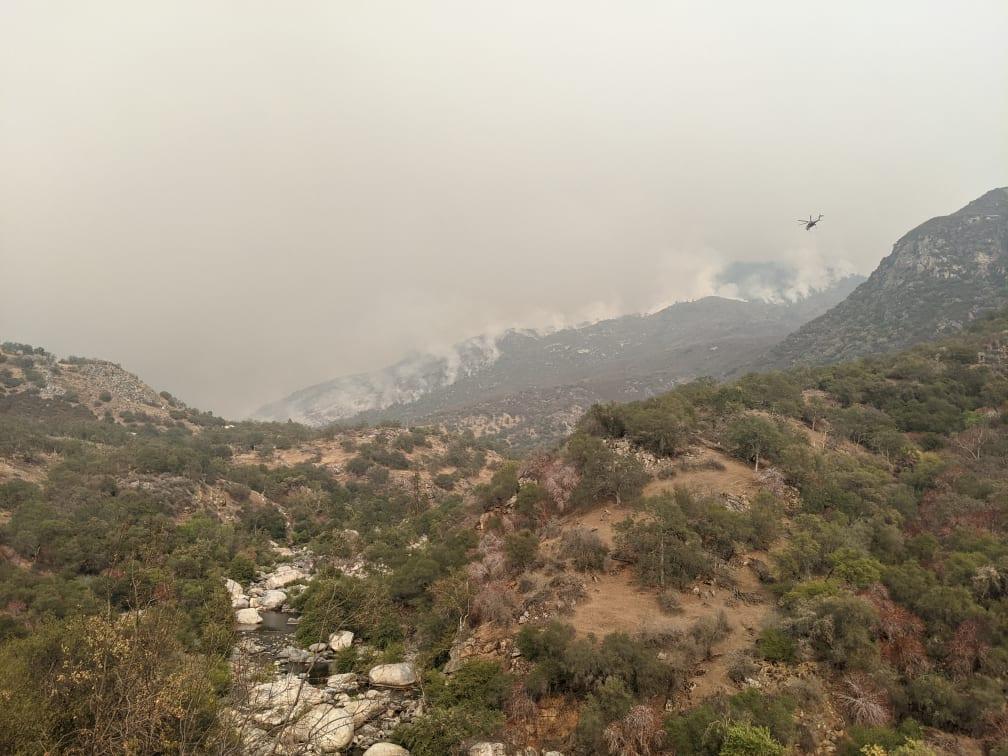 A line of smoke rises from a forested mountainside with a helicopter flying by and a river below. 