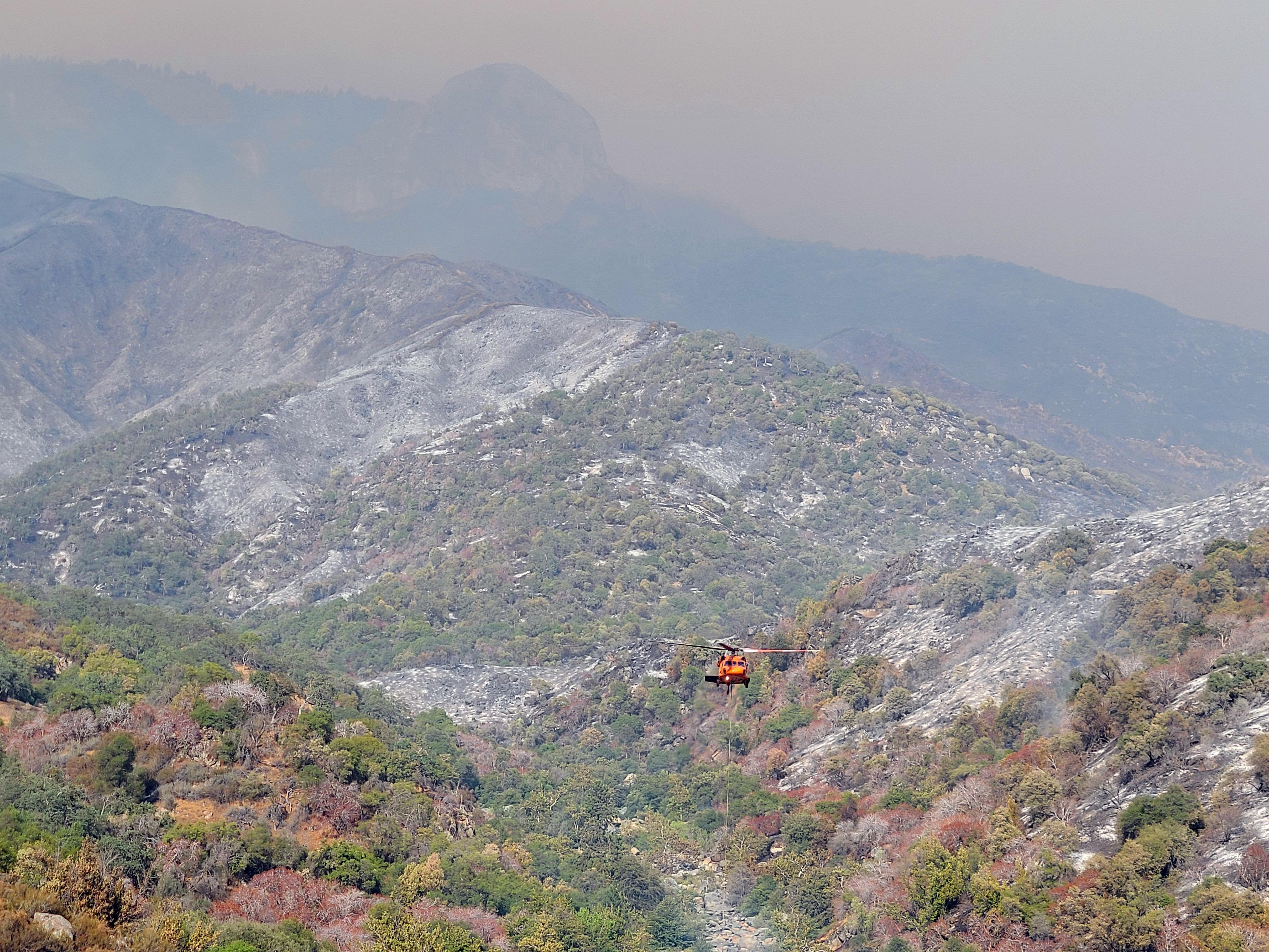 A helicopter flies over a rugged mountain landscape hazed in smoke. 