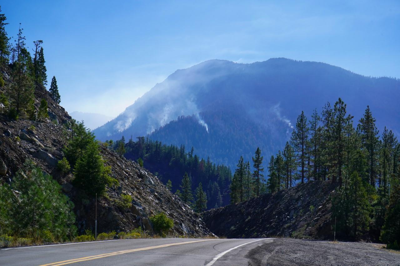 Smoke plumes rising into blue skies from forested mountain with paved road in the foreground.