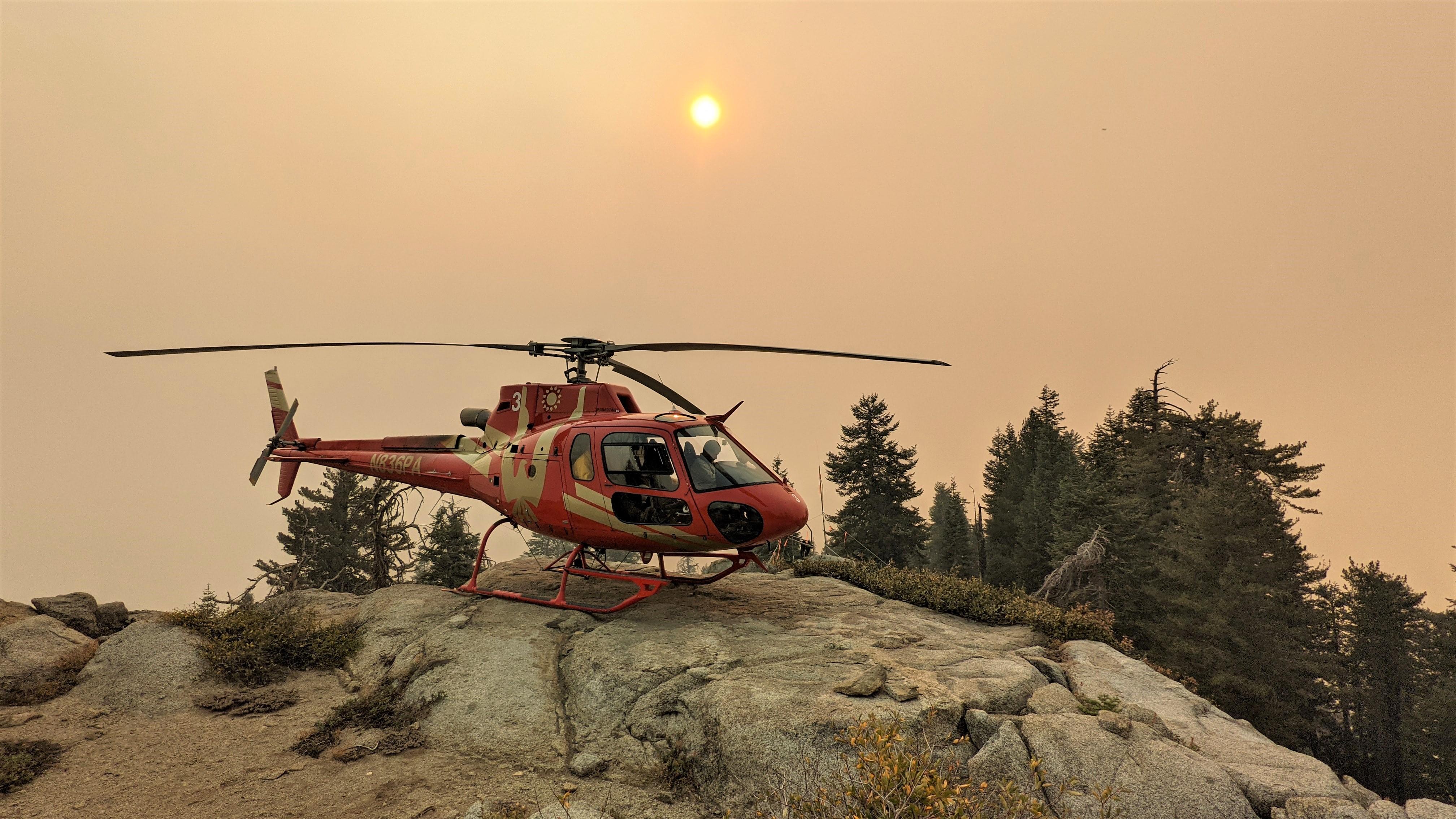 A red helicopter sits on rocks with a smoky sky and red sun