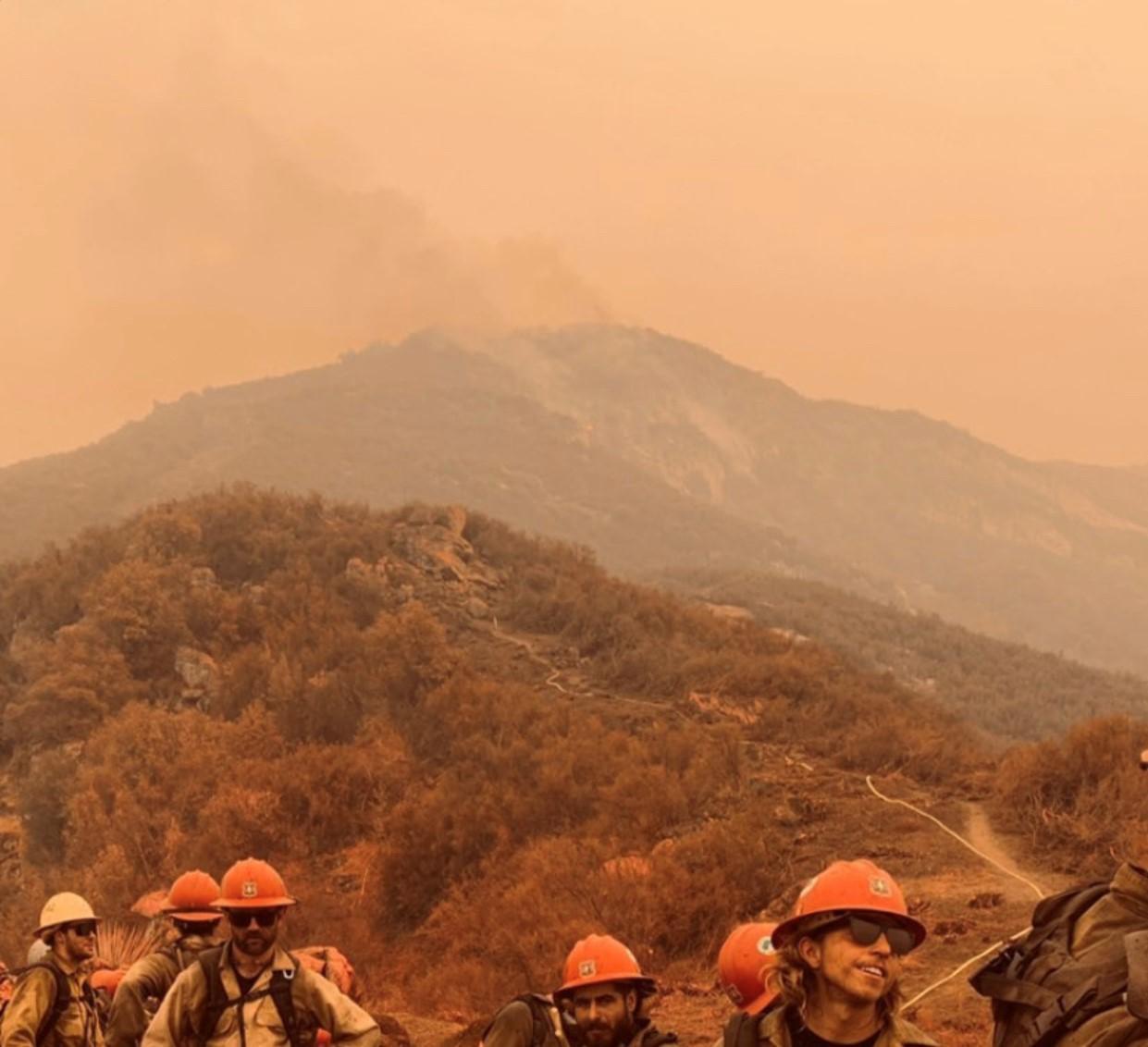 A crew stands in line on a smoky day in front of a mountain with a hose weaving up the hillside along a trail