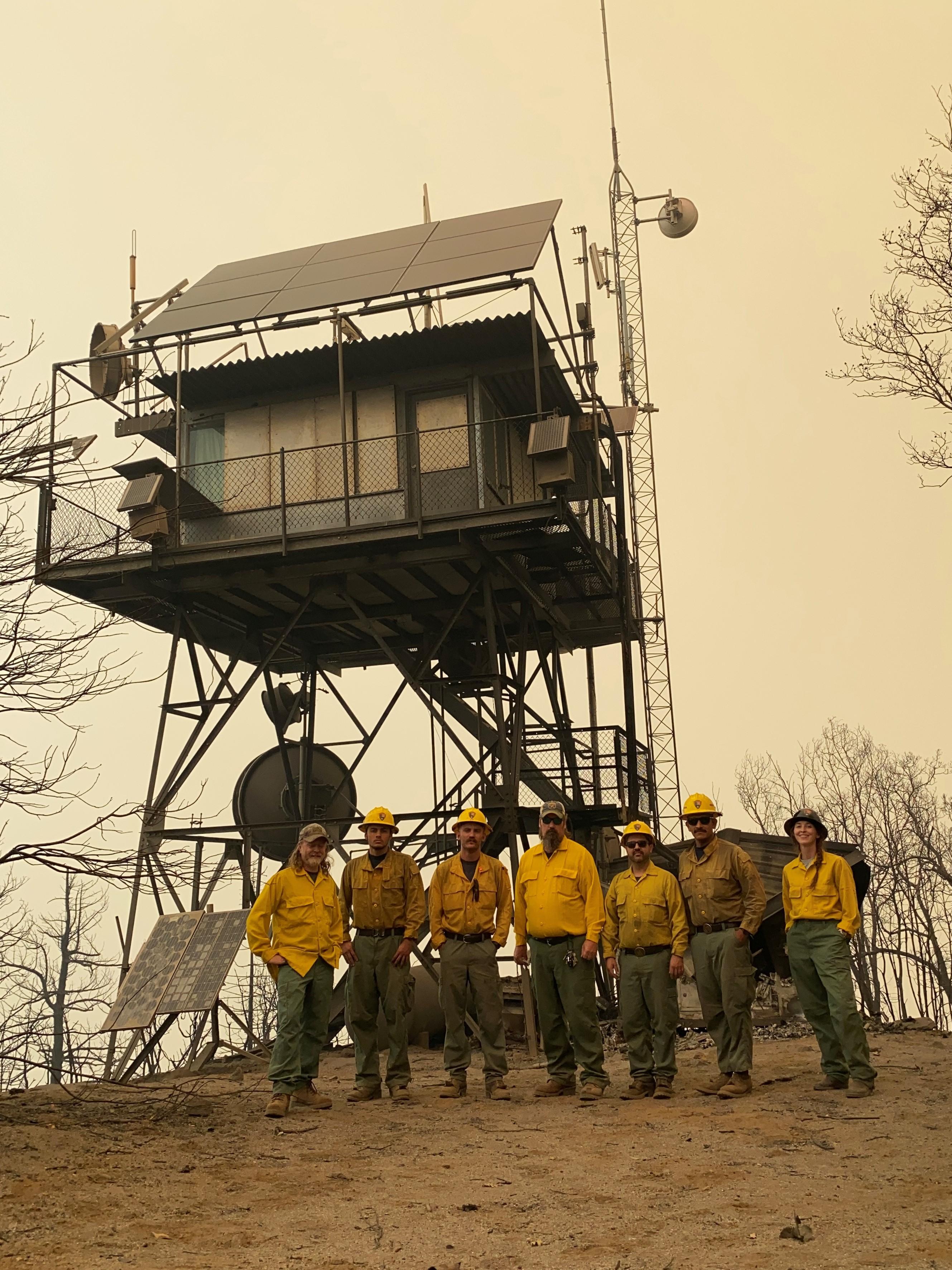 A crew stands below a radio and fire tower on a smoky day