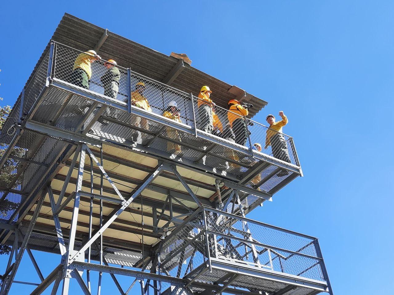 10 people wearing yellow and green clothes are standing on a catwalk of a metal fire tower