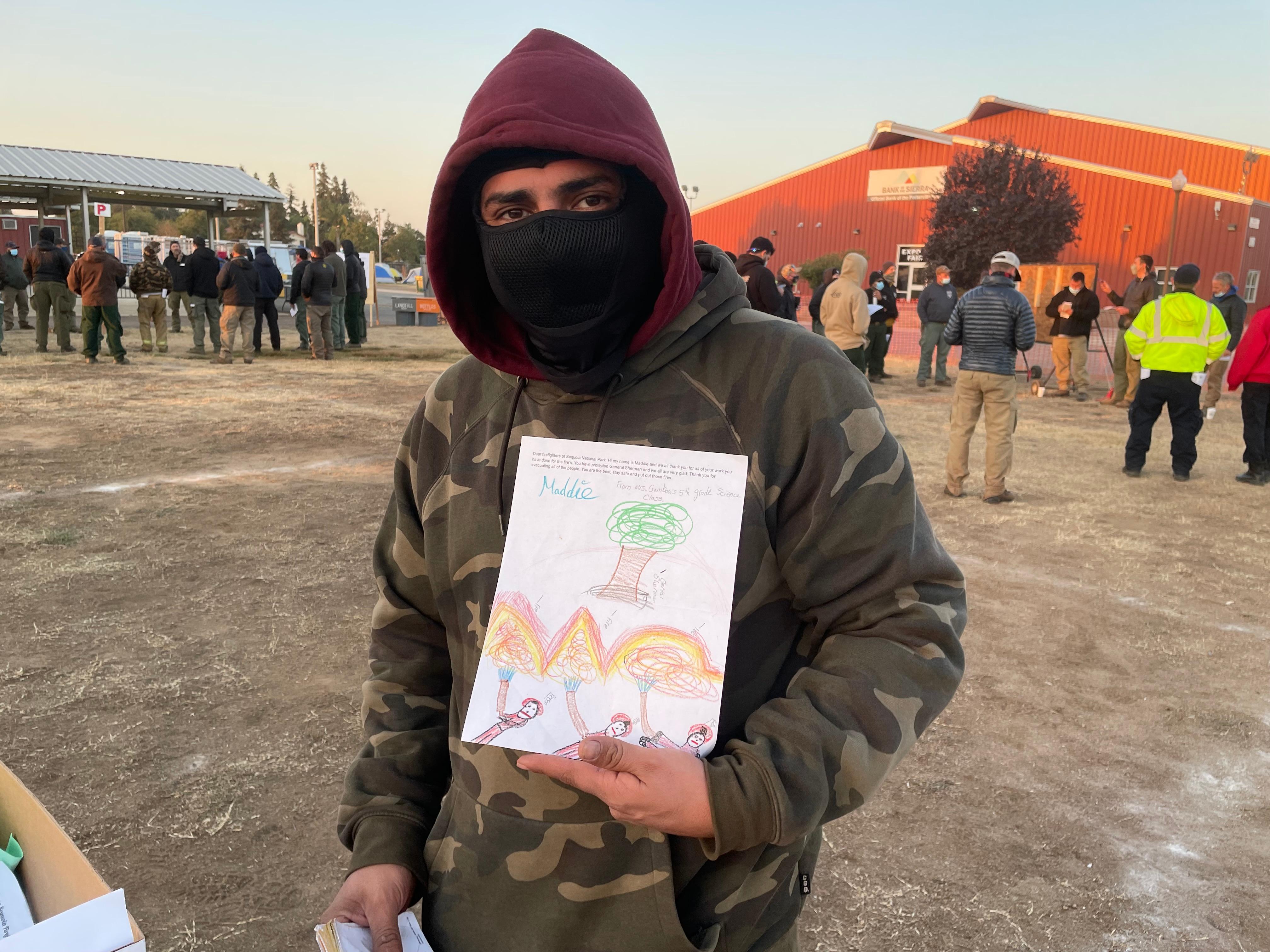 A man in  hoodie holds a letter with a student's drawing of firefighters spraying water on fire to protect a tree. In the background, other firefighters are in groups discussing their work assignments for the day.
