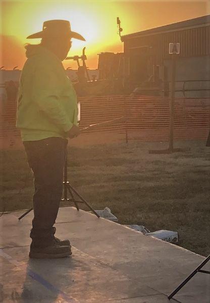 Joey Garfield stands on a stage, backlit by the rising sun. He holds a ceremonial clapstick in his right hand.