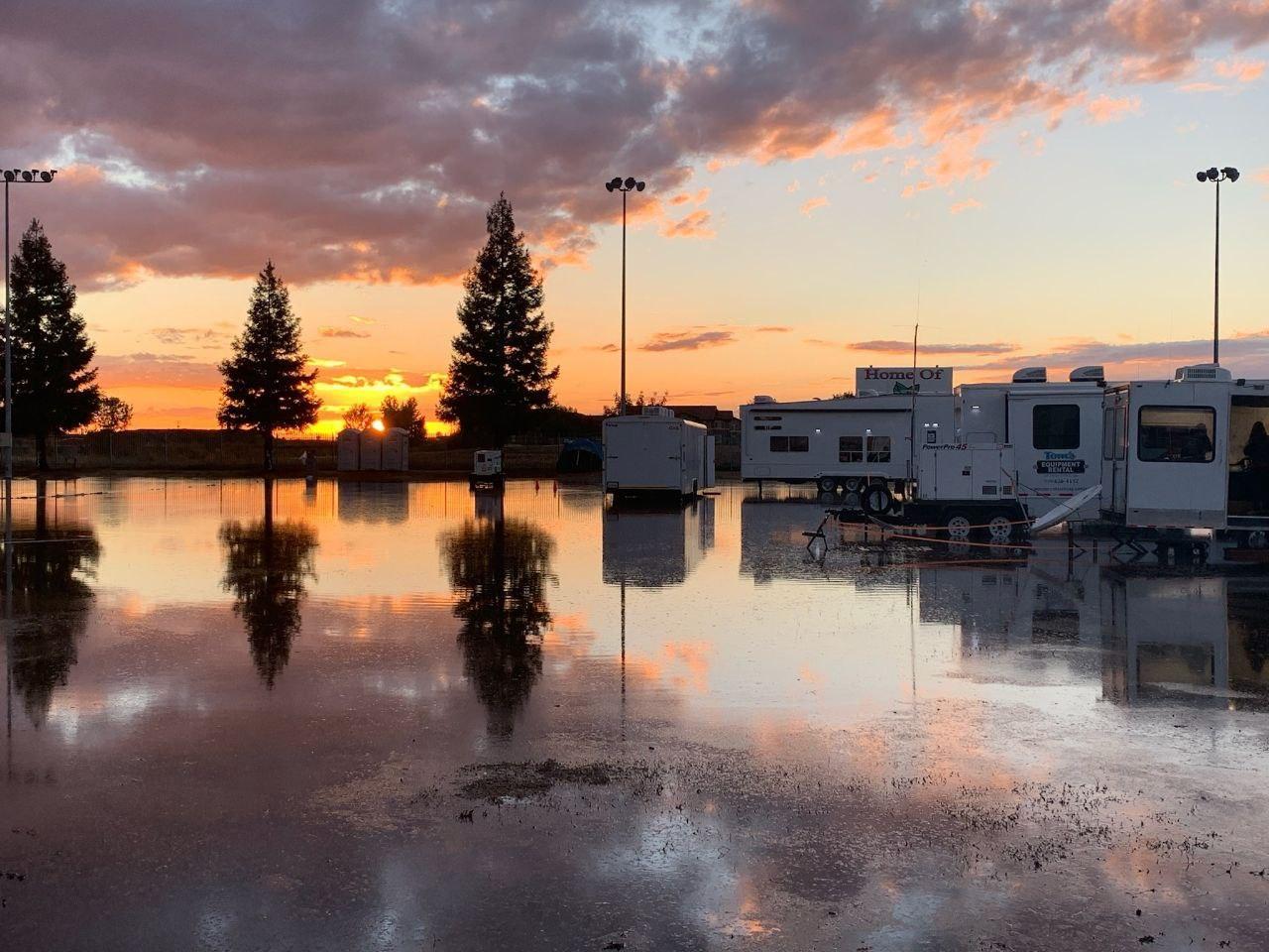 Multiple white travel trailers surrounded by water as sun sets in background.  Reflection of sunset and 3 trees in water