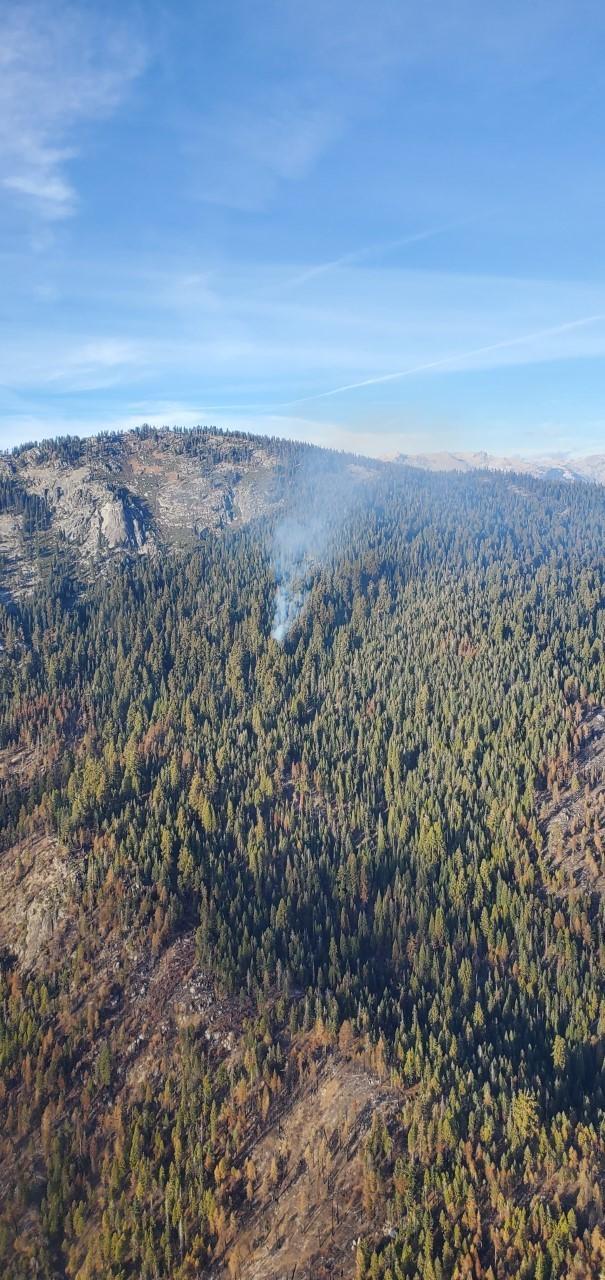Light smoke rises from a forested area below a rocky peak.
