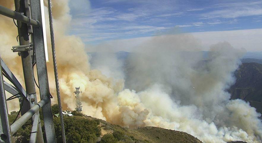Large amounts of smoke rises from a chaparral-covered hillside. The photo is taken from a communications tower.