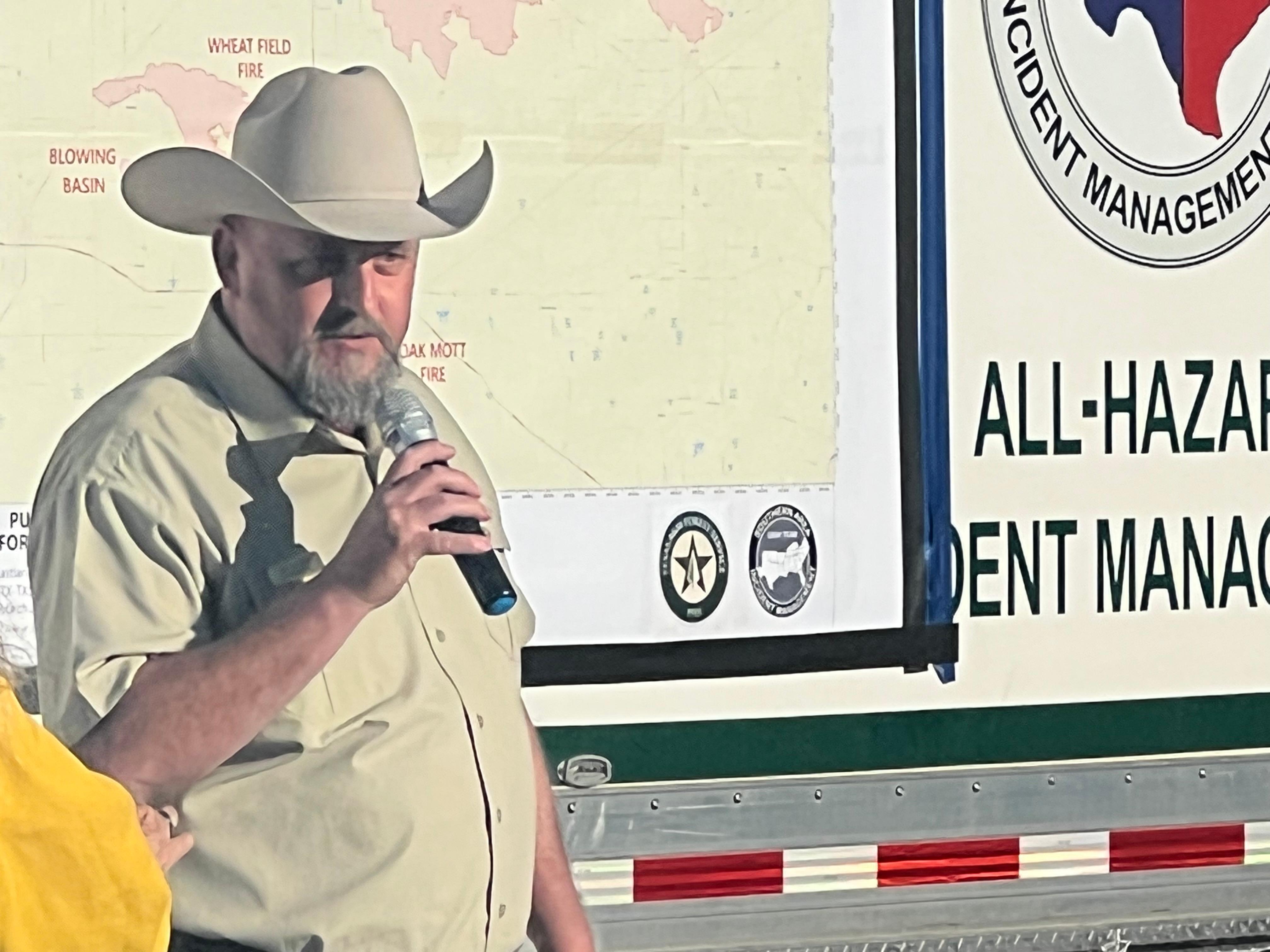 In this picture there is a gentleman wearing a Texas A&M Forest Service uniform and a cowboy hat, speaking to the attendees at the community meeting