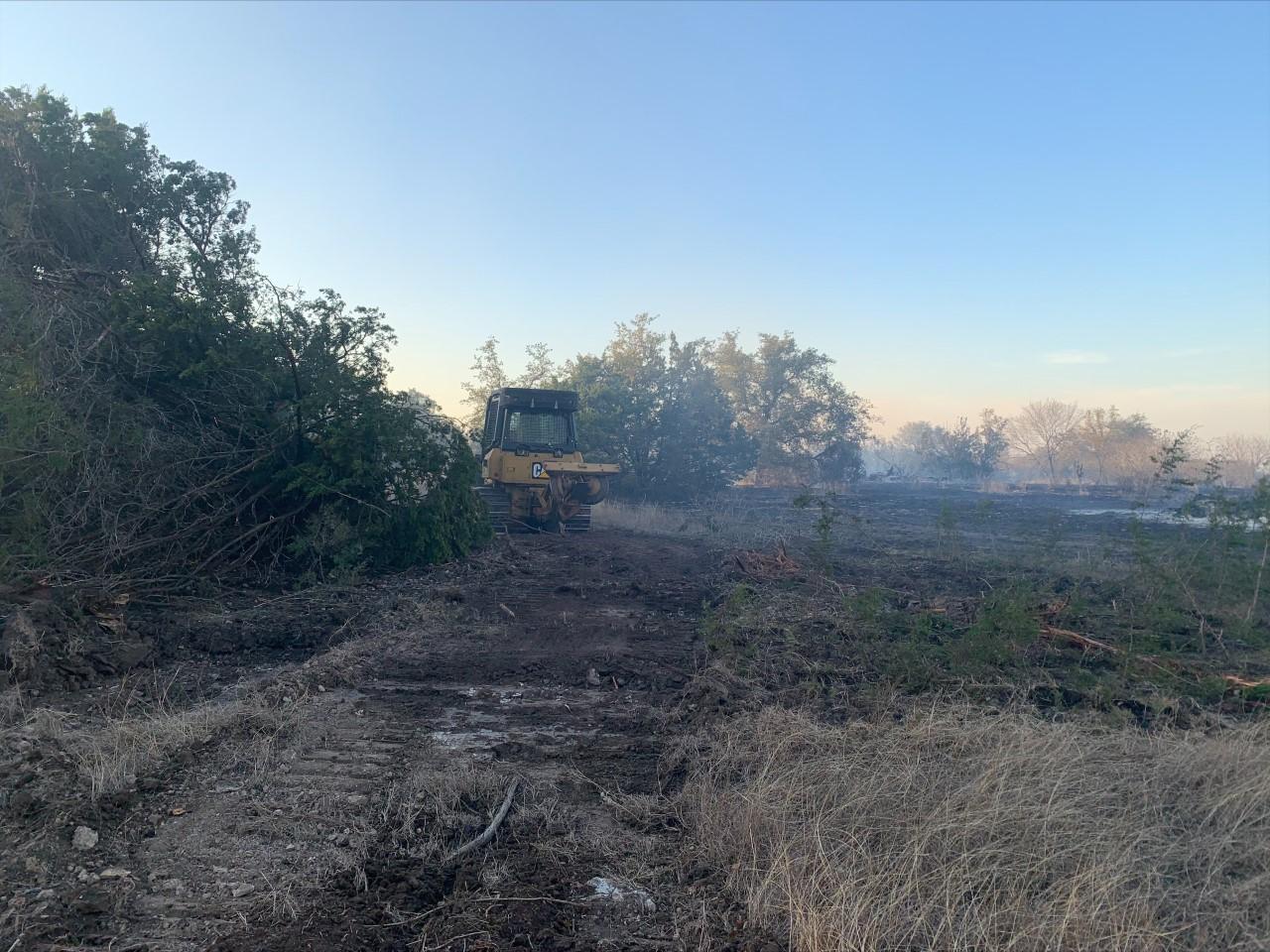 Bulldozer pushing containment line with burned fuels to the right of the line and unburned trees and shrubs to the left