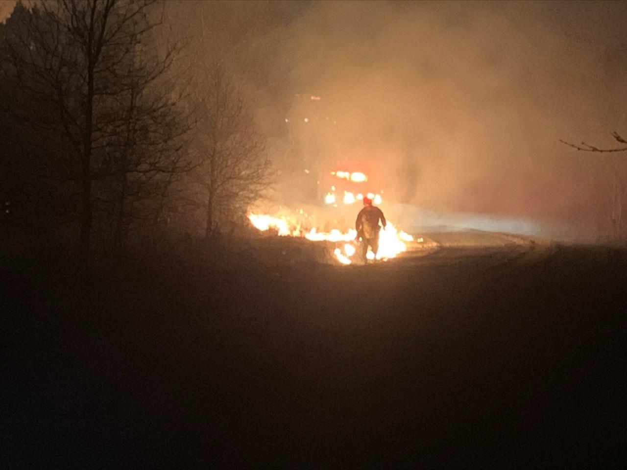 Firefighter walking a dark road with a drip torch lighting a backing fire to create containment line. Glow of engine lights in background