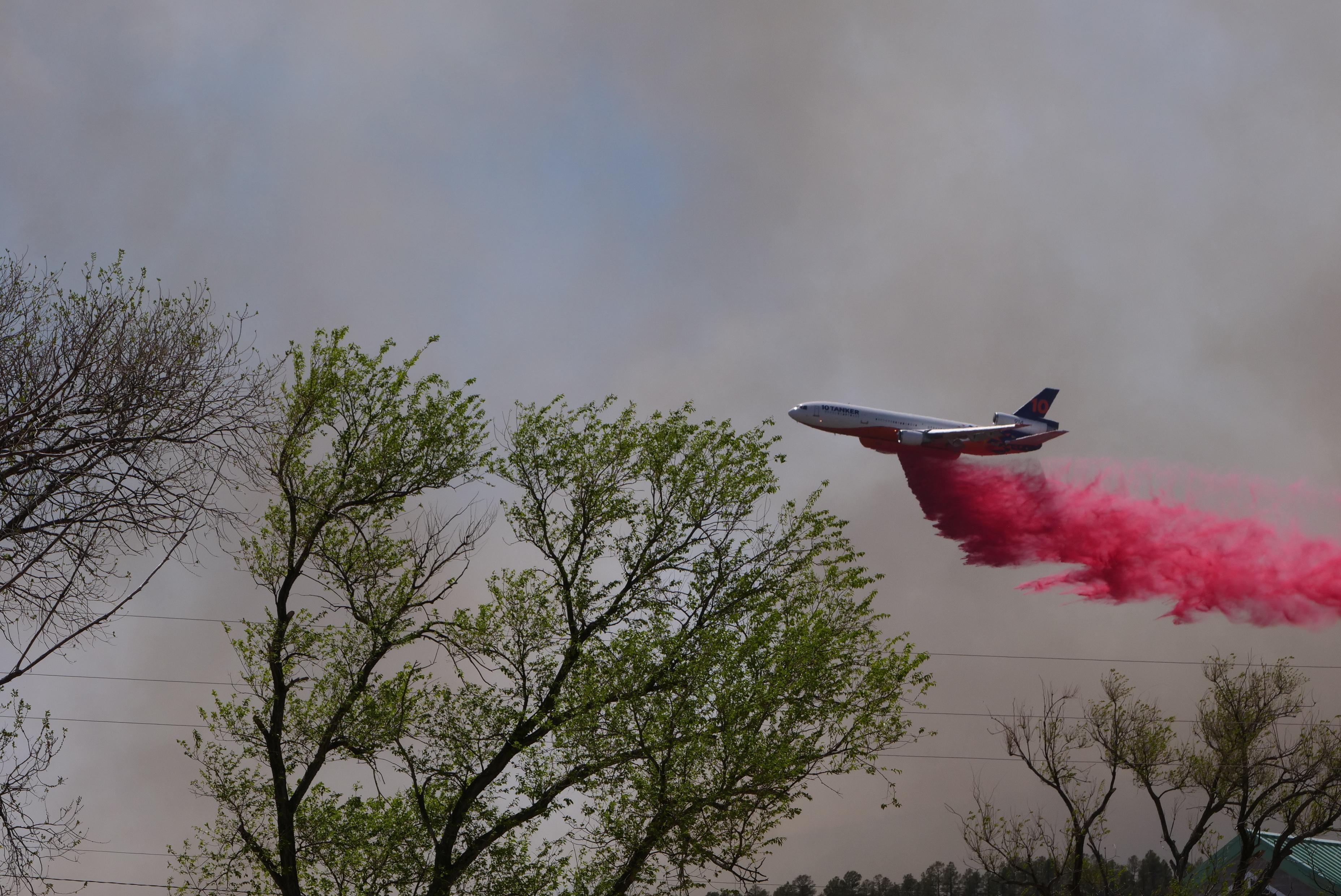A large aircraft is shown releasing red colored fire retardant. Trees are visible in the foreground.