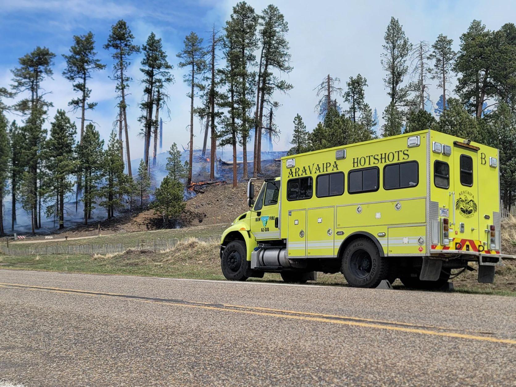 A crew transport vehicle in parked along a road with smoke and fire on a hill in the background