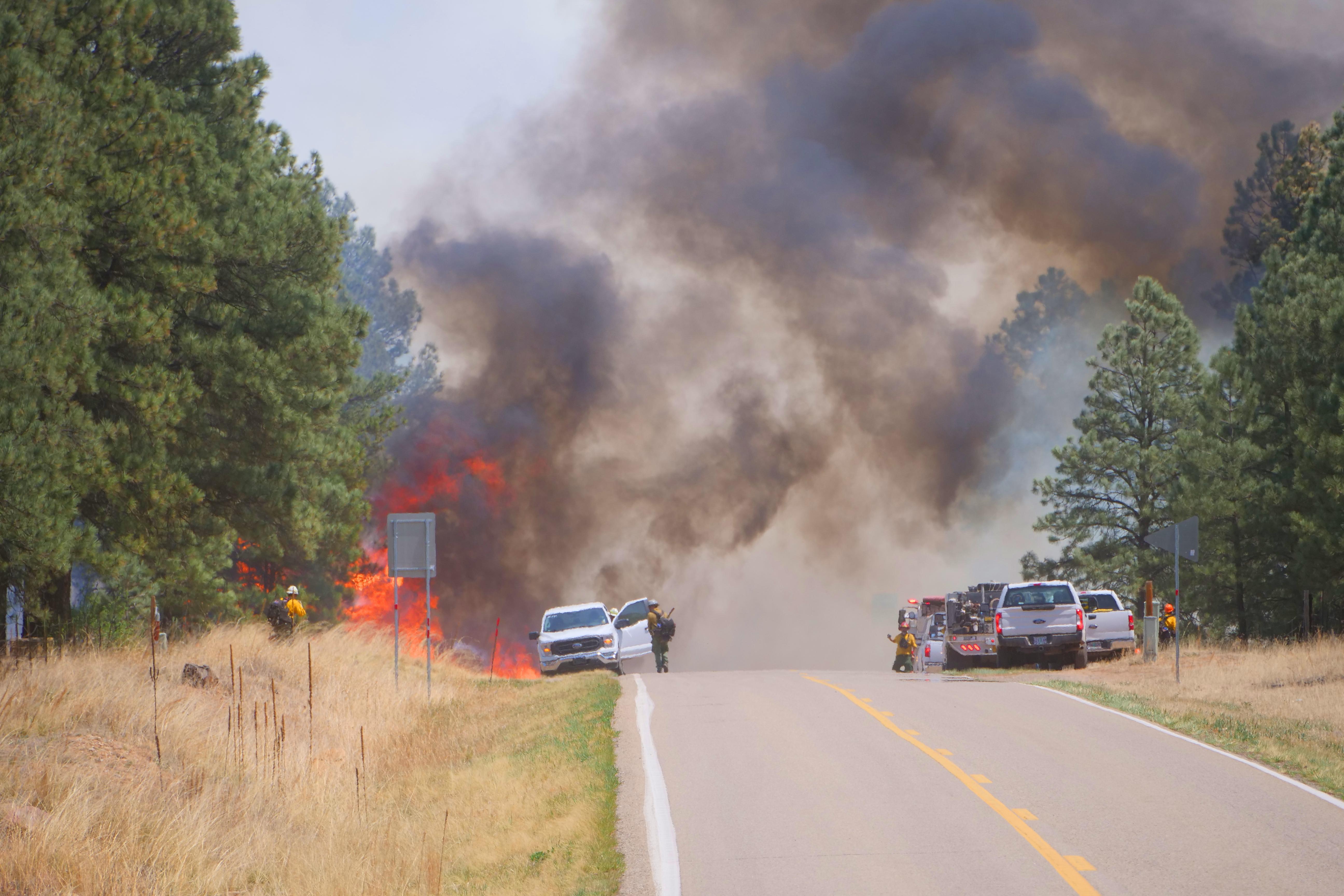 A paved highway is shown in the foreground with flames coming out of trees in the distance. Firefighters and fire trucks can be seen along the road.