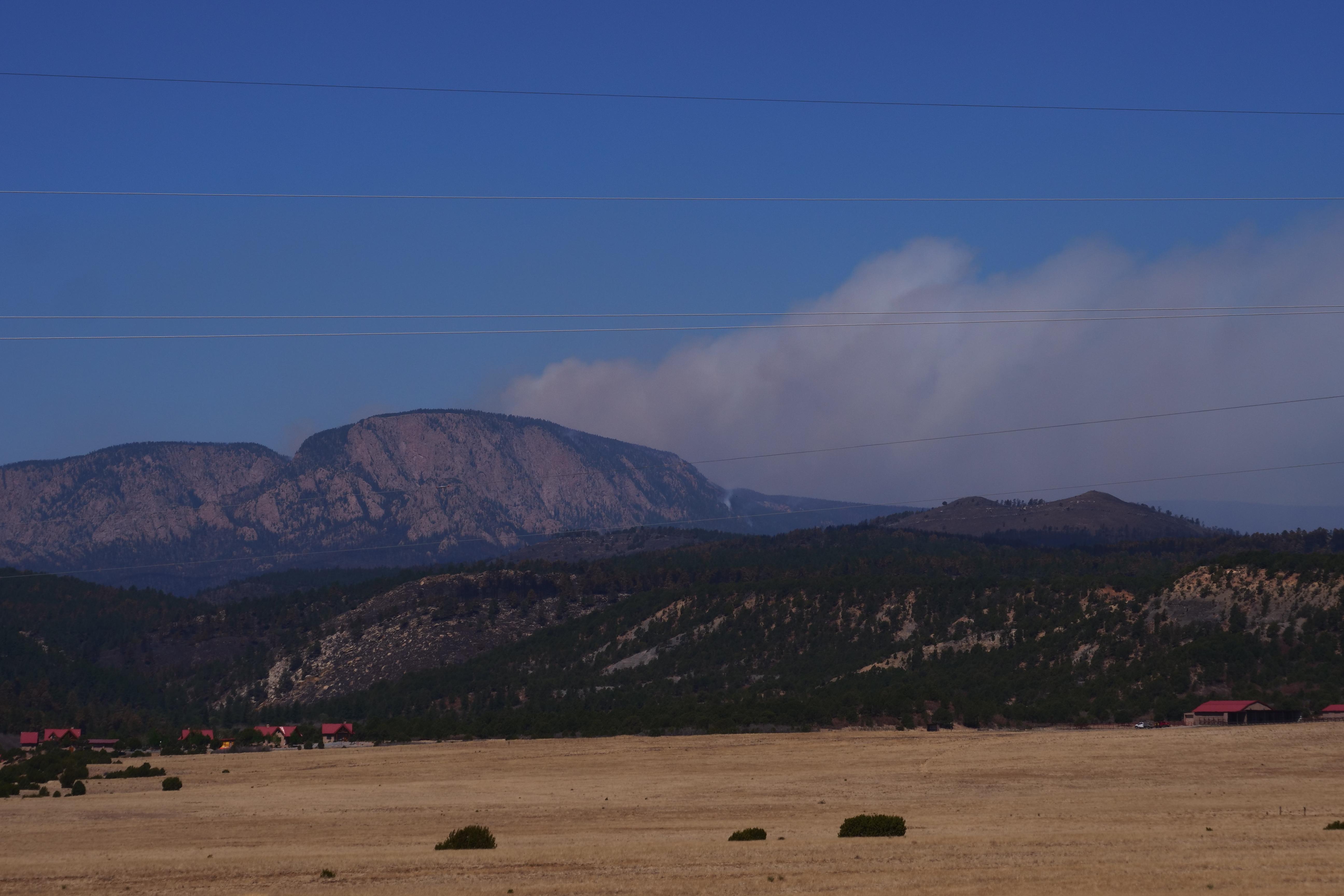 A mountain named Hermits Peak is shown in the distance with a column of smoke visible behind it.