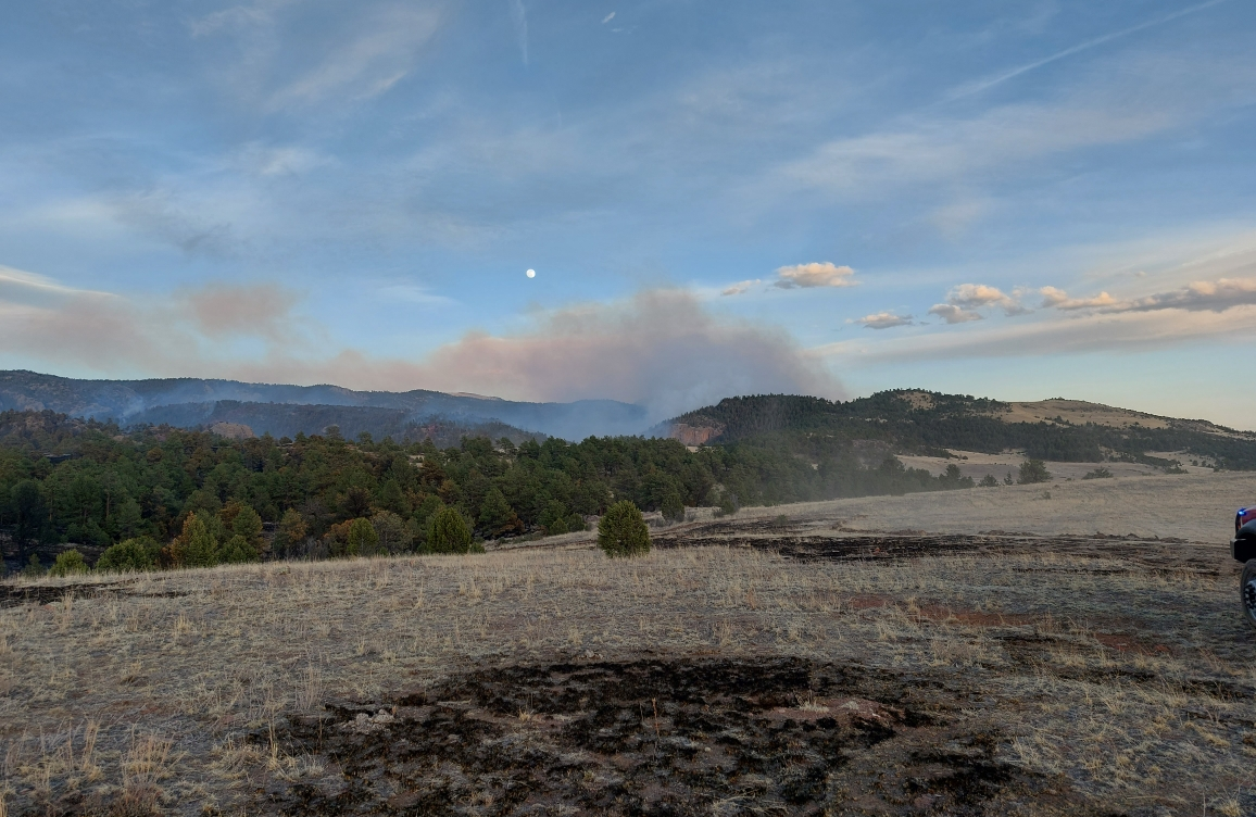 Smoke settles in the valley as evening temperatures cool. Charred grass is visible in the foreground.