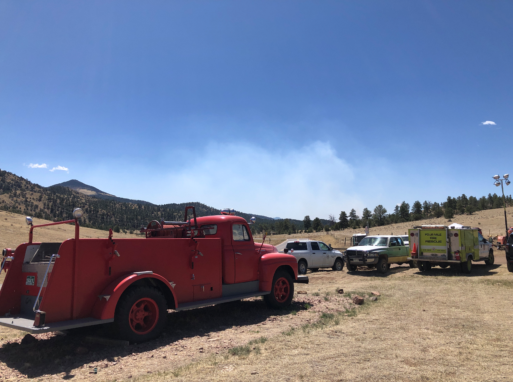 light smoke rises over the horizon from the south end of High Park Fire. An old firetruck is in the foreground, and modern vehicles in the lot.