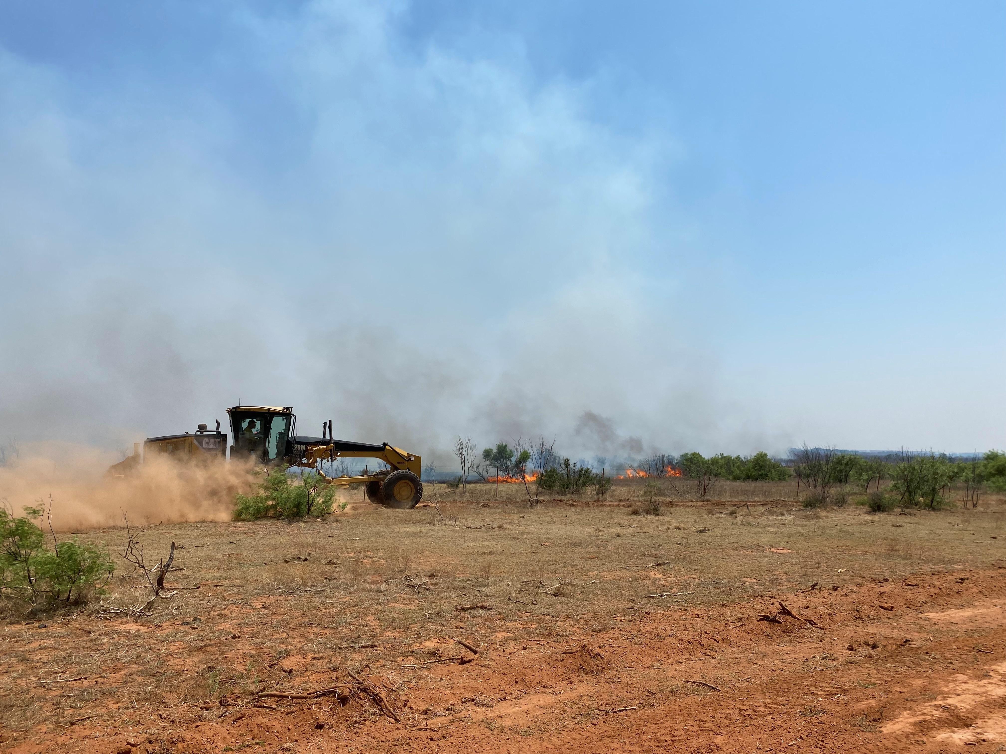 in this picture you can see a motor grader moving towards the right with its blade down widening an already stablished dozer line. in the background you can see smoke and fire coming towards the dozer line.