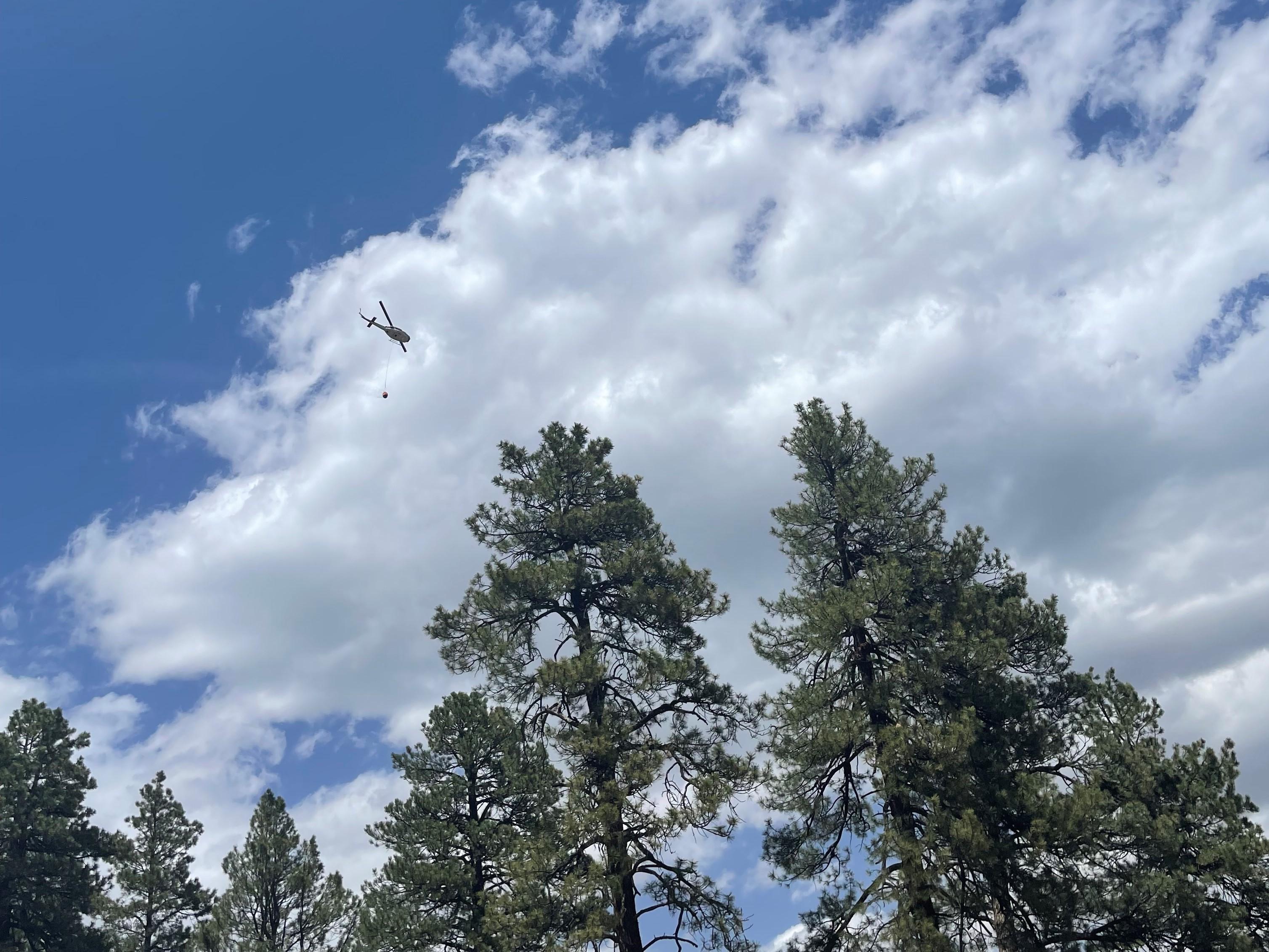 Blue sky, trees, and cloud dominate the view. A medium-heavy helicopter carries a full bucket of water.
