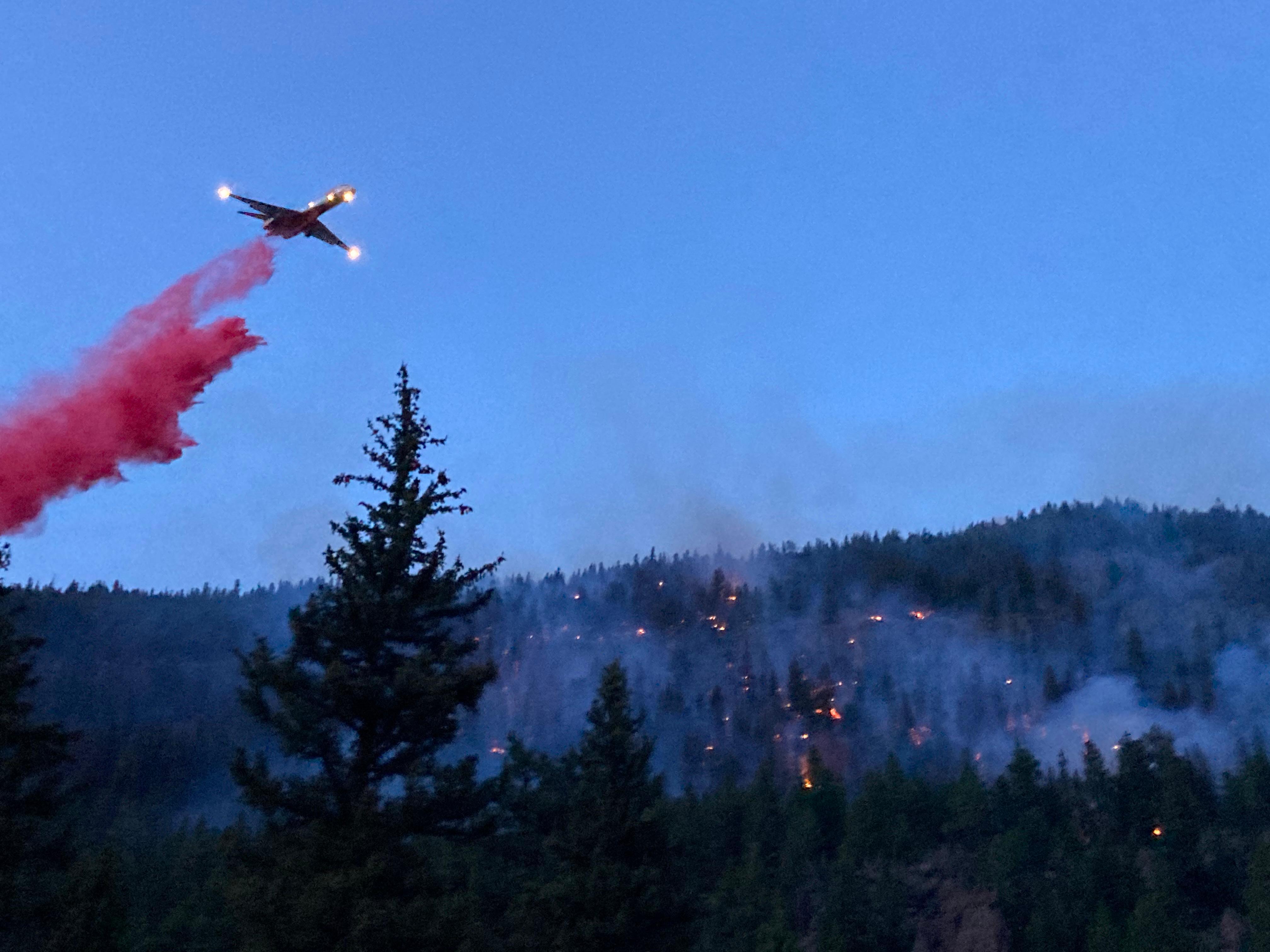 A large airtanker with headlights on drops red fire retardant out over a forest that is speckled with spot fires in the forest.