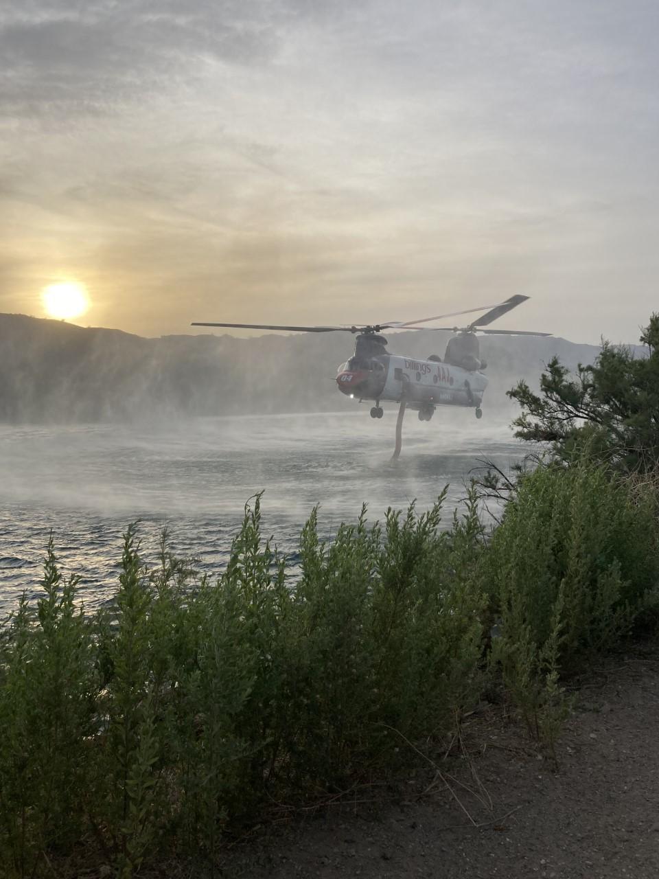 Large helicopter hovers over river, green grass in foreground, river in background