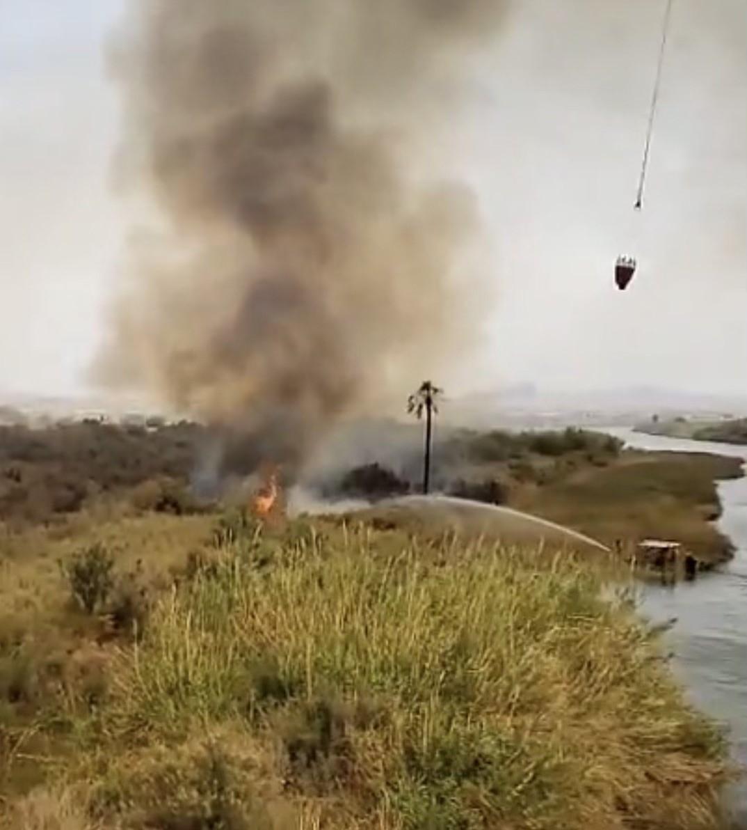 Flames burns salt cedar brush. A boat near river's edge sprays water while a bucket from a helicopter drops water on fire