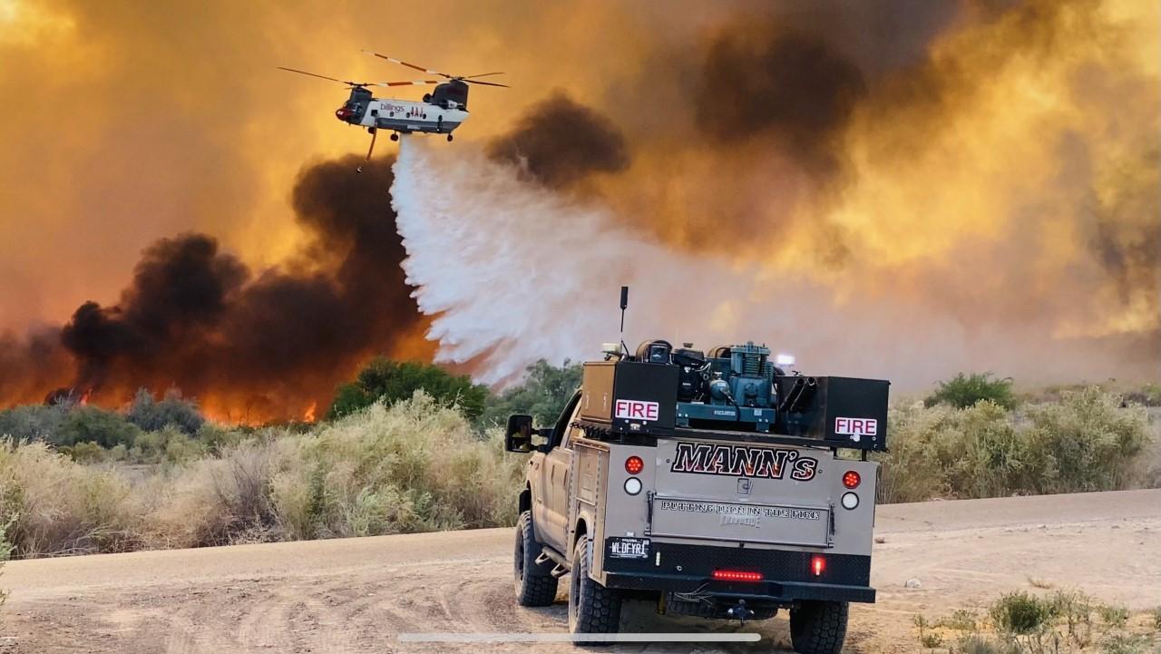 A helicopter drops water over flames with an engine in the foreground