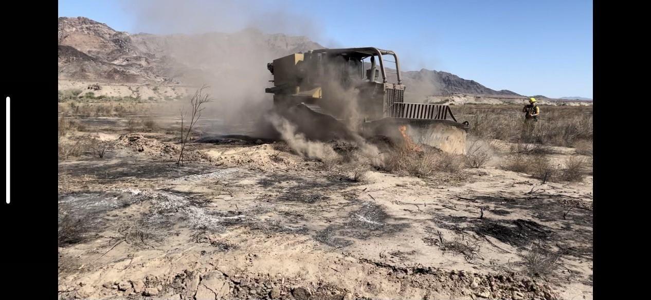 A bulldozer moves tan dirt that has been burned. Firefighter stands to the right of picture talking on radio.
