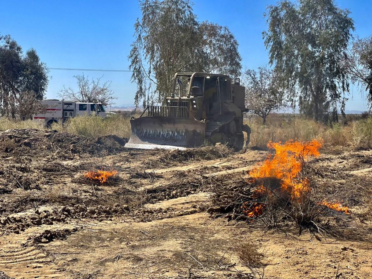 A bulldozer spreads a burning pile of woody debris and dirt apart