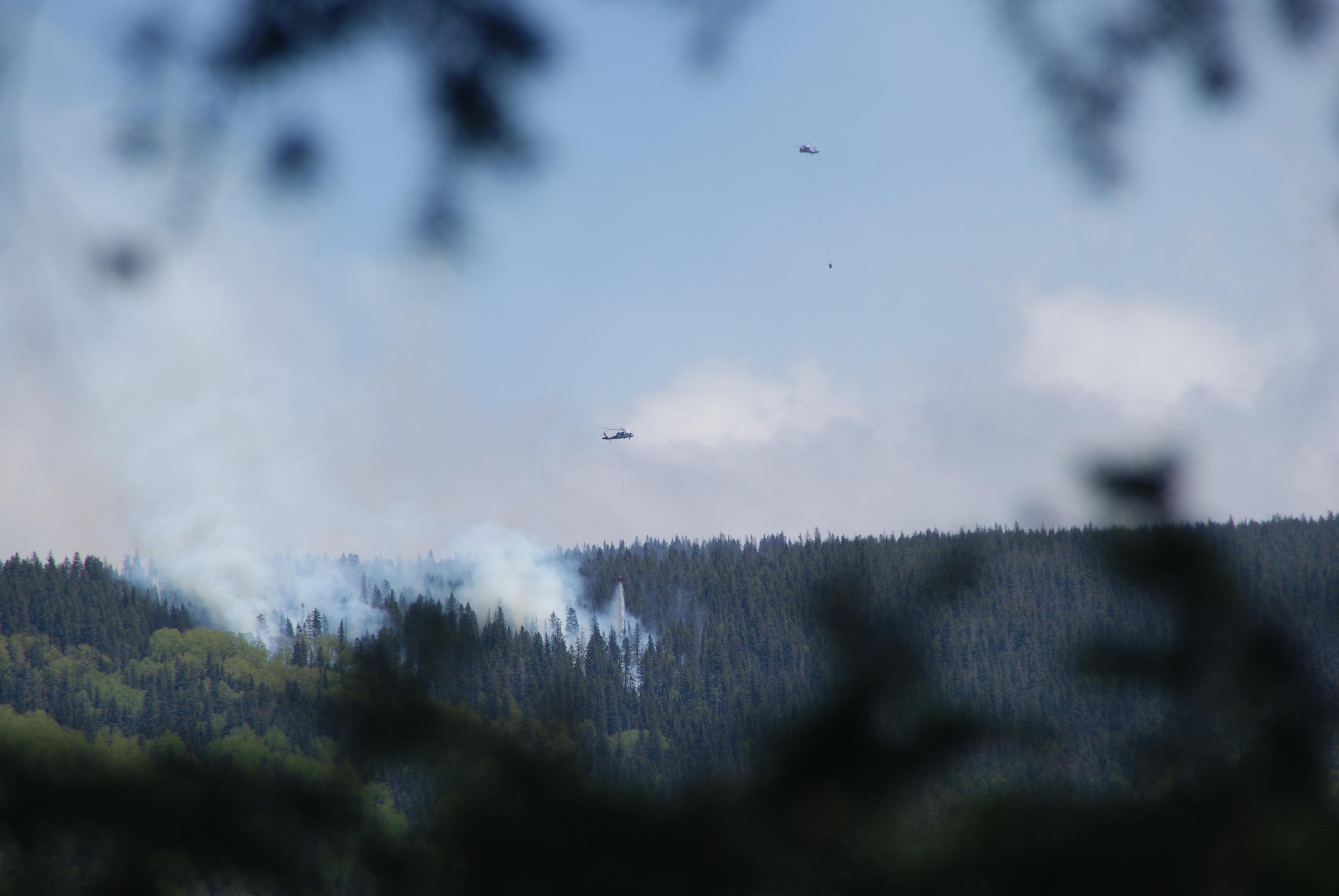 2 helicopters fly over heavily forested terrain. Each helicopter has a long line with a bucket attached to carry water and drop on fire as smoke rises from the trees.
