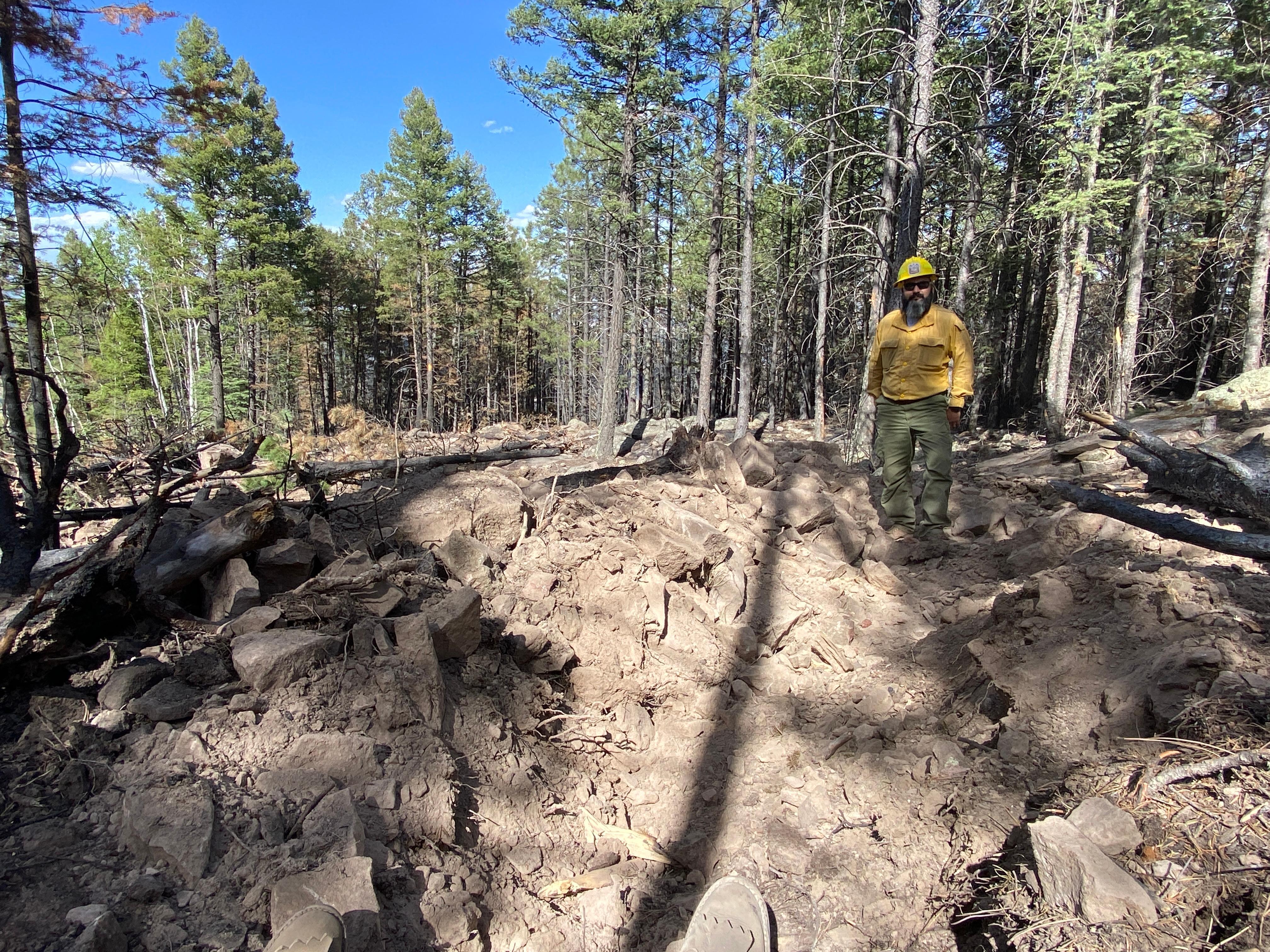 Firefighter in yellow shirt and hardhat stands in a constructed water bar built up with rocks.  Rocks and trees covering a previously constructed dozer line.