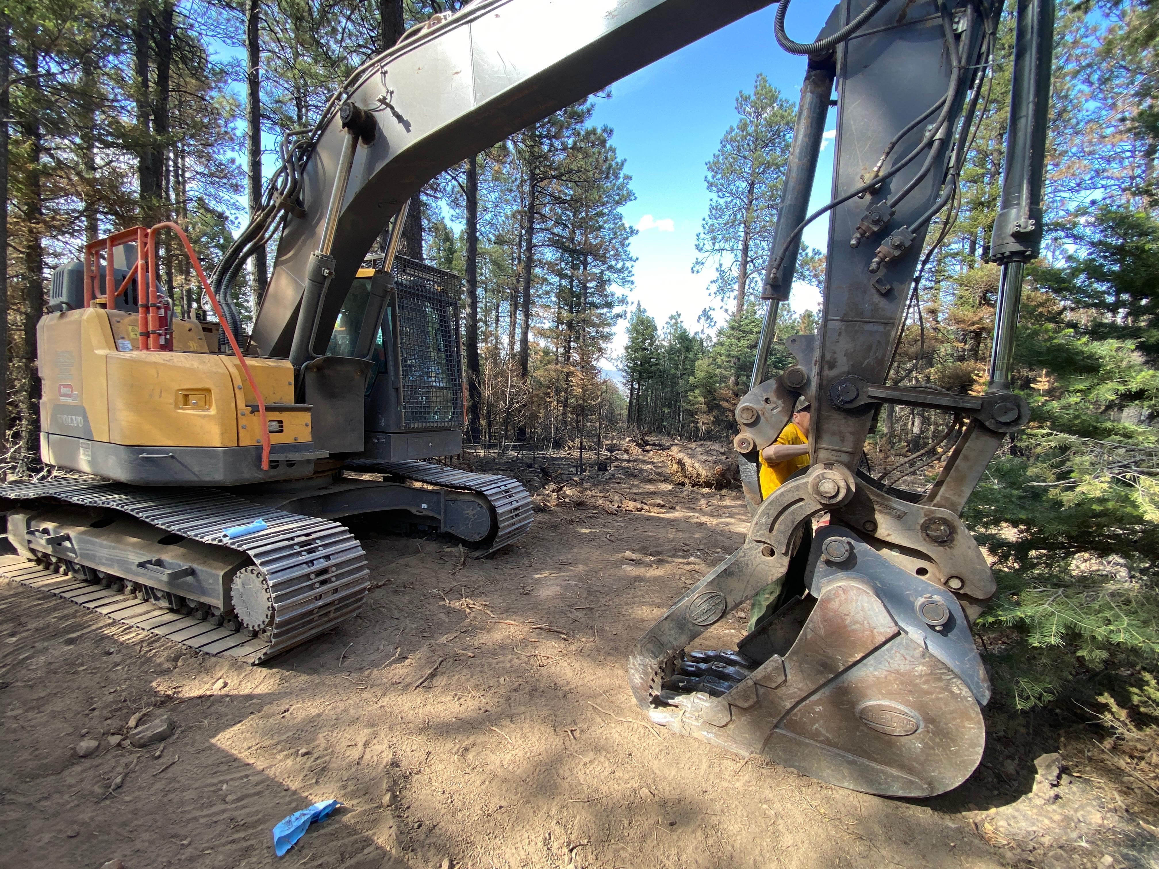 Image of an excavator, heavy piece of equipment. Machine is black and yellow with a bucket on the end of an arm extension. Surrounding area is a mix of burned and unburned vegetation. Heavy equipment operator in yellow shirt is standing behind equipment