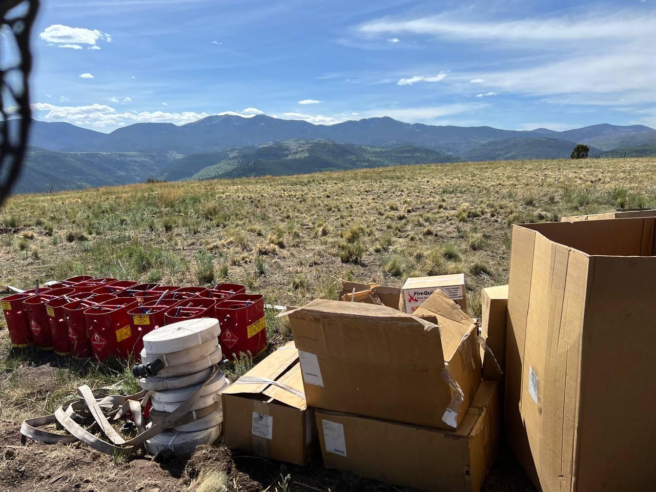 Multiple red gas cans, rolls of white hose, and cardboard boxes of equipment sit in an open meadow