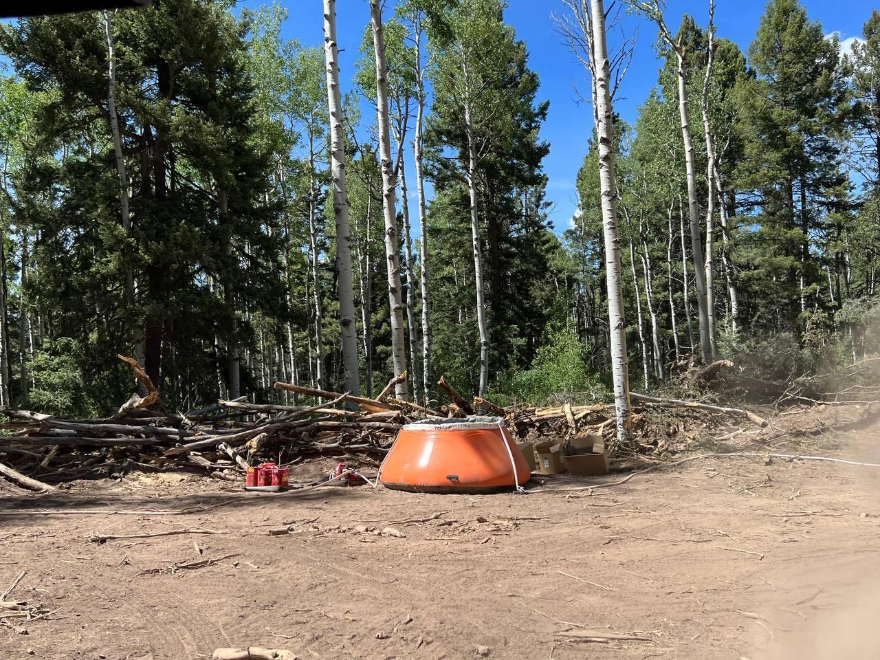 Orange tank known as a pumpkin with white hose, red gas cans, red pump sitting on a cleared area of dirt.  Pile of downed trees behind equipment with forested area in background.