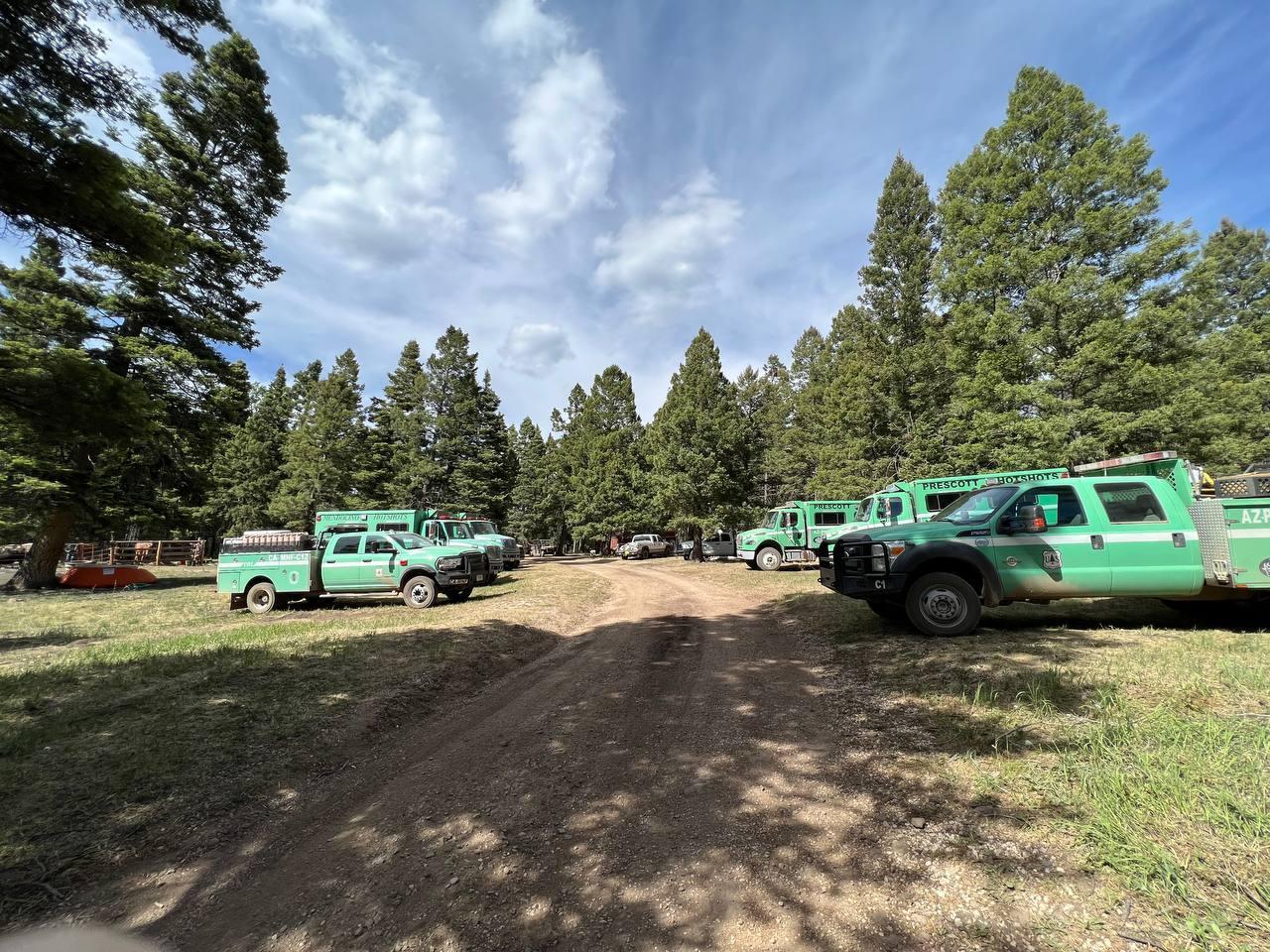 Green fire trucks and hotshot crew buggies parked in campground with trees and cloudy, blue sky in background