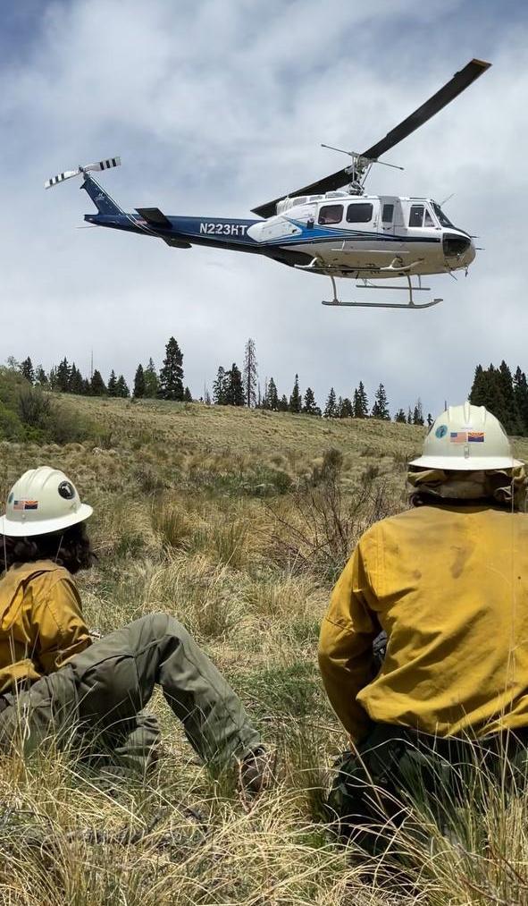 Two firefighters with yellow shirts, green pants, and hardhats sit in meadow. White and Blue helicopter lifting off from ground against a cloudy, blue sky
