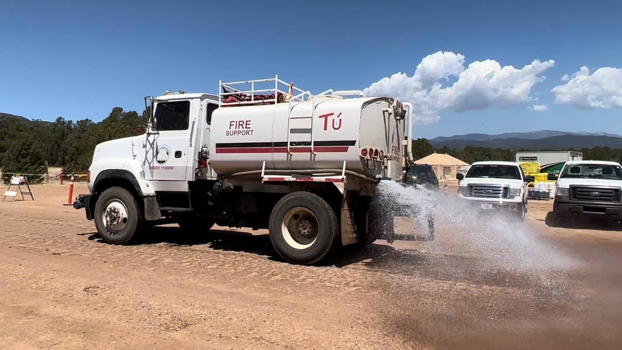 Large white truck with water tank on the back driving on dirt road. Water is spraying from the rear of truck and onto ground to prevent dust blowing. Vehicles and cloud, blue sky in background