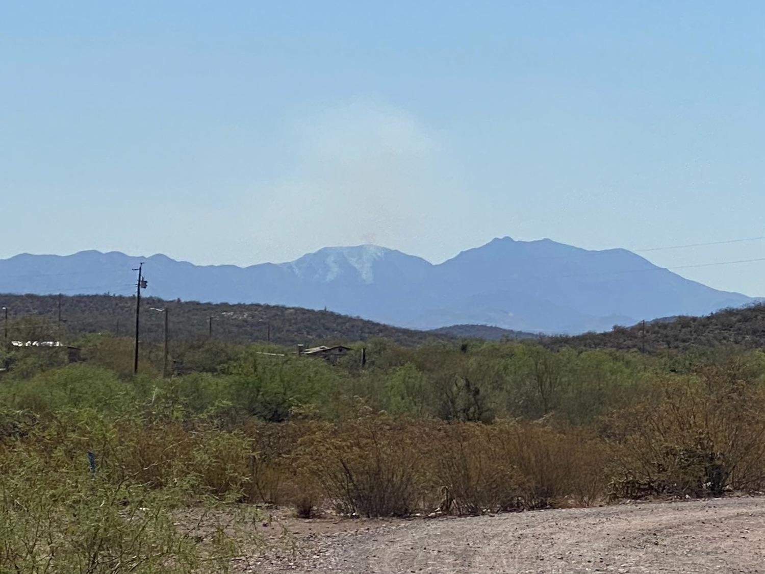 A house is surrounded by desert vegetation with smoke coming up from center of picture in the background.