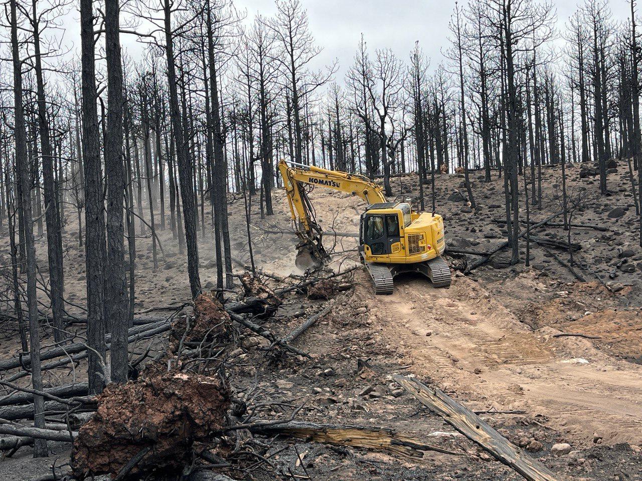Piece of Yellow, heavy equipment called an excavator working to drag burned trees over dozer line.  Slope has many standing, burned trees across the landscape.