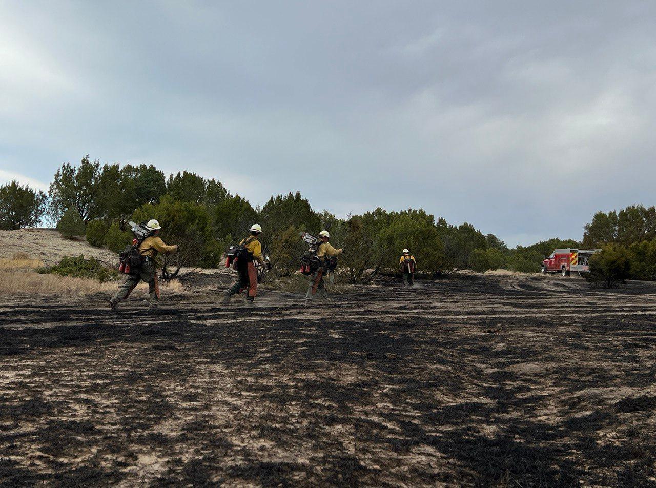 4 firefighters walk across burned, black ground with vegetation and a red engine truck in the background.  2 firefighters are carrying chainsaws.