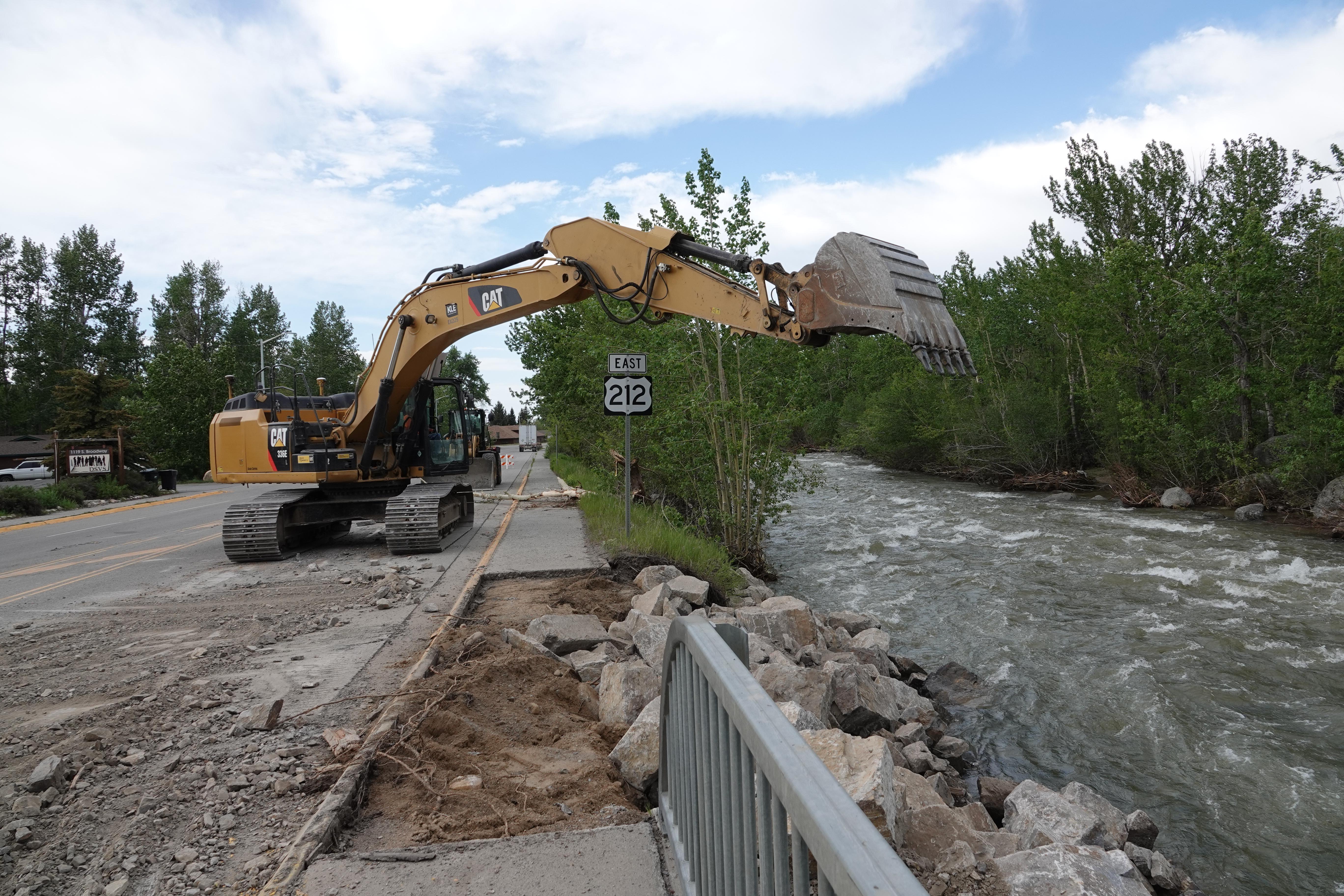 Heavy equipment with bucket in the air on US-212, creek with large rocks, damaged highway edge