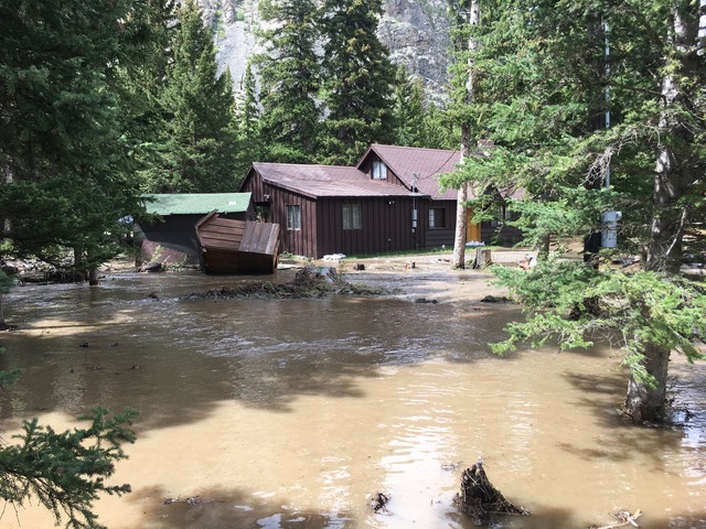 Damaged cabin in background, muddy flood waters in foreground
