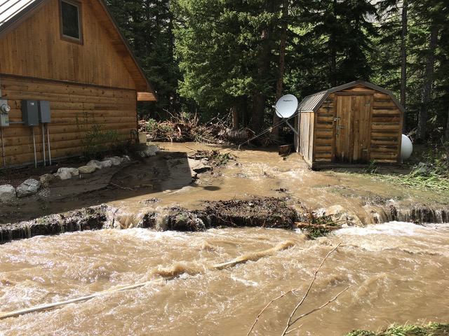 Muddy flood waters flowing between a log cabin and a log shed