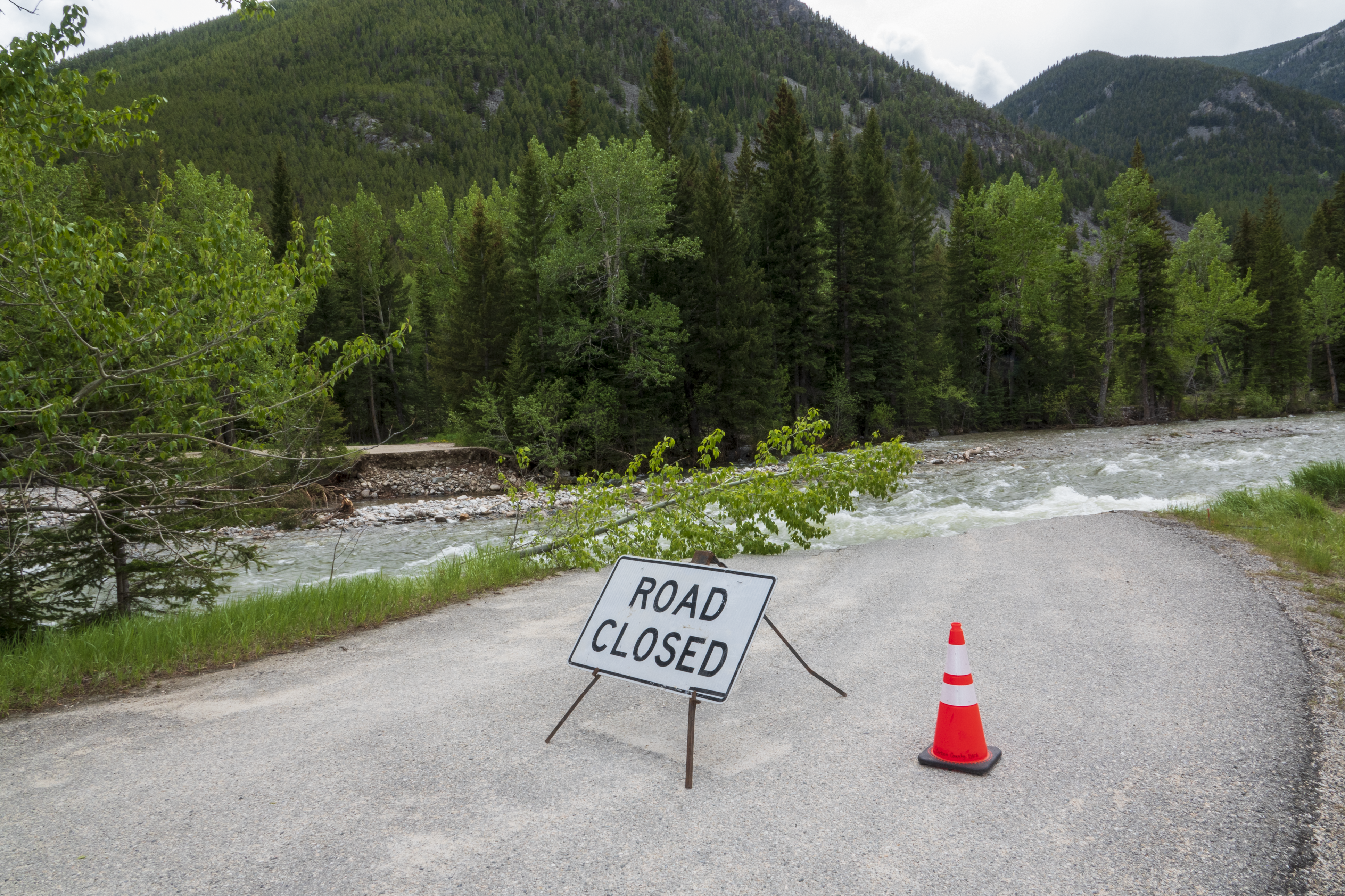Road closed sign and single orange cone on pavement, broken pavement along both sides of creek running to left of image
